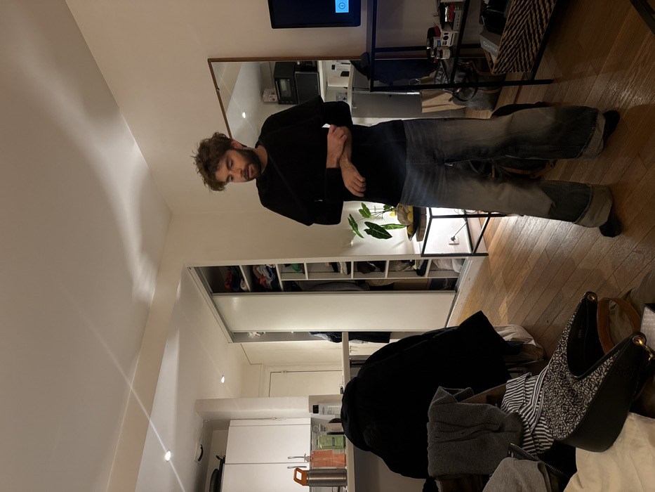 A man stands in an interior with light hardwood floors. He wears a black long-sleeve shirt and faded blue jeans. A large mirror hangs on the wall behind him, reflecting a kitchen area. In the foreground is a pile of clothes and a black handbag. A white wardrobe with sliding doors is to the left of the man. A small table holding a houseplant is next to him. A television is mounted on the wall above the mirror.