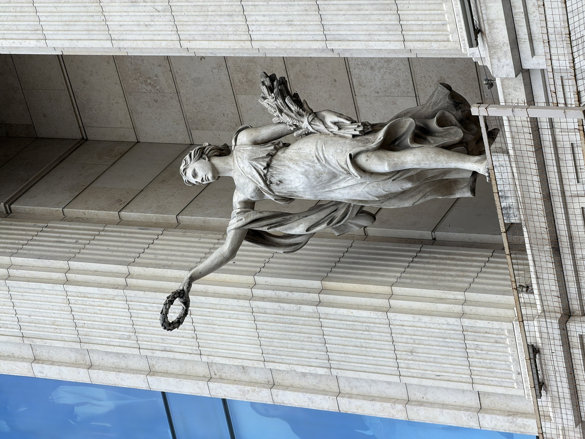The image shows a marble statue of a female figure, likely representing a goddess or an allegorical personification, situated on the facade of a building. The statue is depicted in a classical style, with flowing robes and a serene expression. She is holding a laurel wreath in her outstretched right hand and a bundle of wheat stalks in her left arm. The statue is positioned in an alcove, framed by fluted columns. To the left of the statue, a large section of the building's facade is covered by a blue glass panel, reflecting a silhouette of a person. The building appears to be constructed of stone blocks with a ribbed texture. In the foreground, a wire fence with metal posts is visible, suggesting a balcony or a protected viewing area. The overall impression is one of grandeur and historical significance. This statue is located on the facade of the Singer House, also known as the Dom Knigi, on Nevsky Prospekt in Saint Petersburg, Russia.