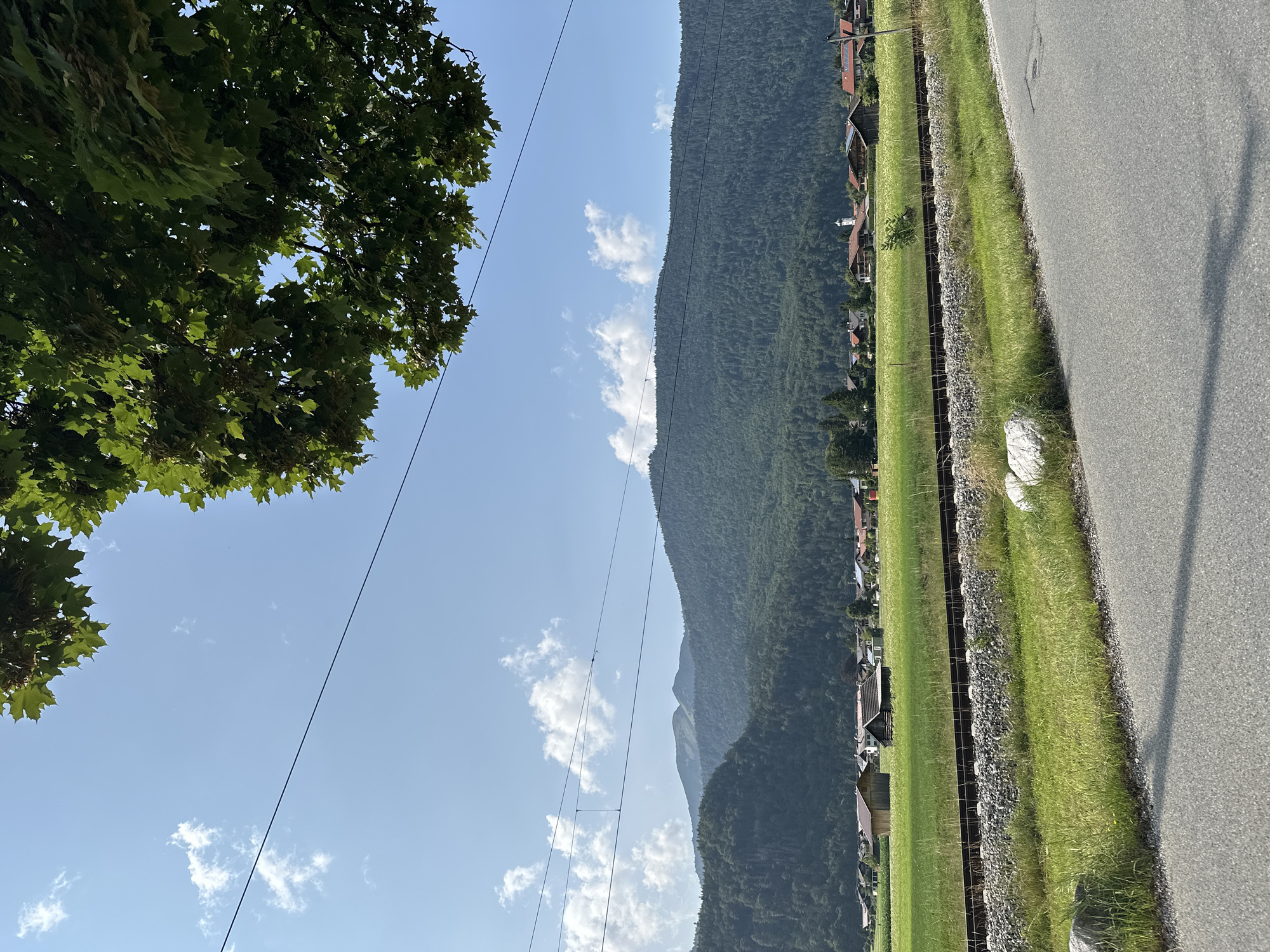 An overcast sky with scattered clouds is visible above a rural landscape. In the foreground, a paved road runs horizontally across the lower portion of the image, with grassy verges on either side. A set of railway tracks with ballast runs parallel to the road, separating the foreground from a lush green field. Beyond the field, a village with traditional Bavarian architecture, featuring wooden chalets with red-tiled roofs, is nestled against a backdrop of rolling, tree-covered hills. A prominent church steeple rises above the village. The distant mountains are covered in dense green forests, and their peaks are partially obscured by haze. The image captures a serene, idyllic scene in Farchant, Germany, bathed in the soft light of a summer day.