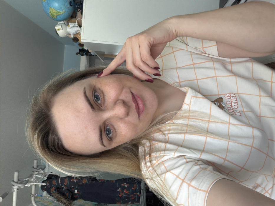 This image depicts a female individual indoors in Votkinsk, Russia, captured from the chest up, looking directly at the camera with a slight smile. She has blonde hair, blue eyes, and dark eyebrows. Her left index finger is positioned against her left temple area, and her fingernails are painted dark red. She is wearing a cream-colored, short-sleeved t-shirt adorned with a light orange grid pattern and a graphic on the chest depicting a brown animal peeking from behind text that reads "THERE IS NO OTHER".
The background indicates an indoor, possibly domestic, environment. To the left, a white multi-level clothes drying rack is visible, from which hangs a dark-colored children's coat or snowsuit featuring a fur-trimmed hood and a pattern of light-brown animals. This is set against a light-colored wall with faint vertical stripes. To the right, a white cabinet or shelf unit displays several objects, including a dark Batman figure, a light-colored astronaut figurine, and a blue globe. The wall behind these objects is a light blue color. The scene is brightly lit.