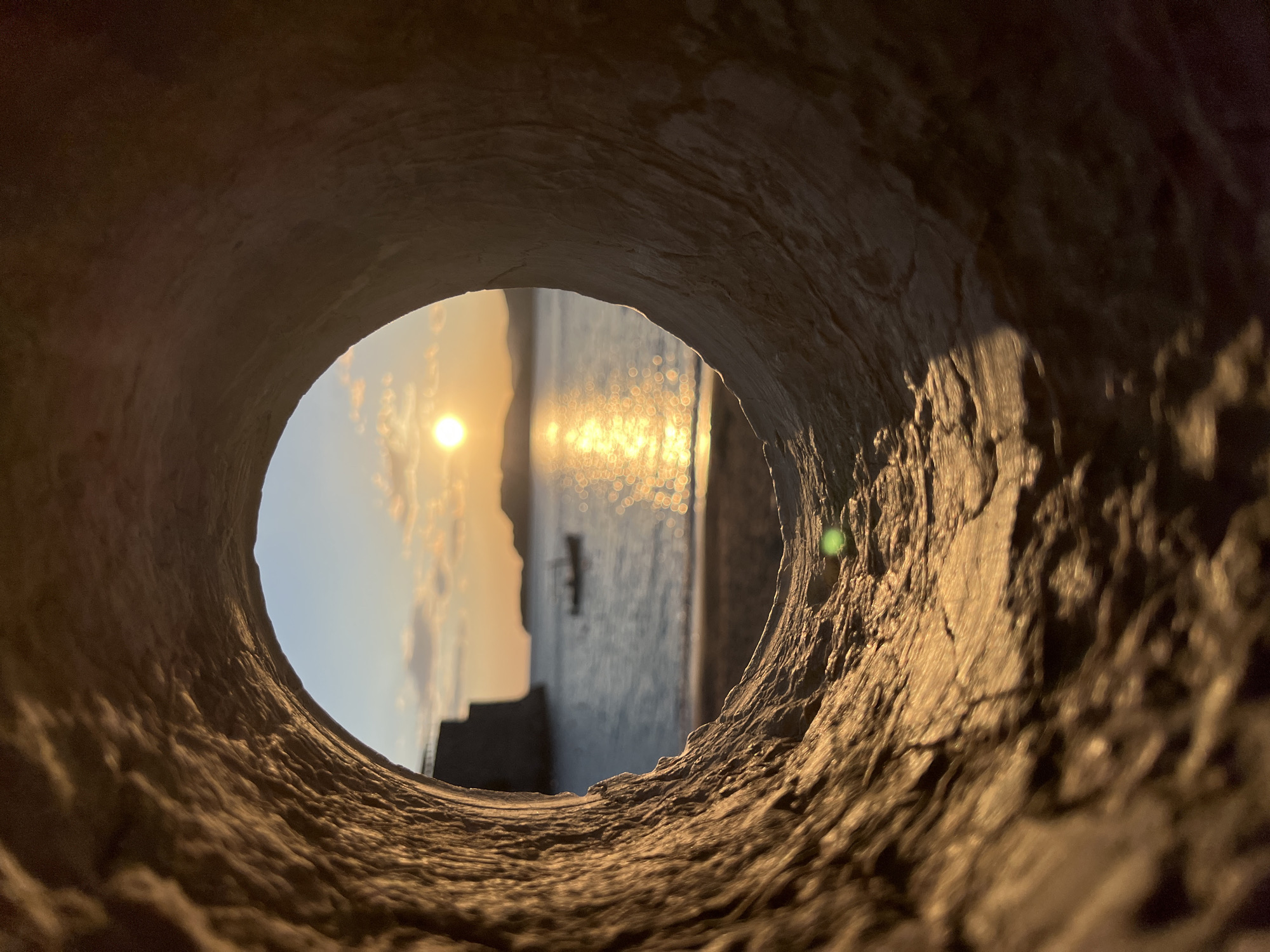 The photograph presents a unique view of a sunrise or sunset, framed by the dark, textured interior of a circular opening, likely a tunnel entrance or a large pipe. The rough, irregular surface of this foreground element is illuminated by warm, golden light, highlighting its tactile details.
Through this framing, a serene scene is revealed: a bright, golden sun, positioned low in the sky, casts its light across the landscape. The sky displays a beautiful gradient, transitioning from soft blue to warm orange and yellow, with a few wispy clouds. Beneath the sky, a body of calm water shimmers with the sun's reflections, creating numerous sparkling bokeh effects across its surface. In the distance, the dark silhouette of a landmass, consistent with the island or coastline of Sveti Stefan, Montenegro, is visible against the horizon. A small, dark boat rests peacefully on the water near the shore.
The image captures the "golden hour" light, evoking a sense of tranquility and a unique perspective. There is no visible text in the image.