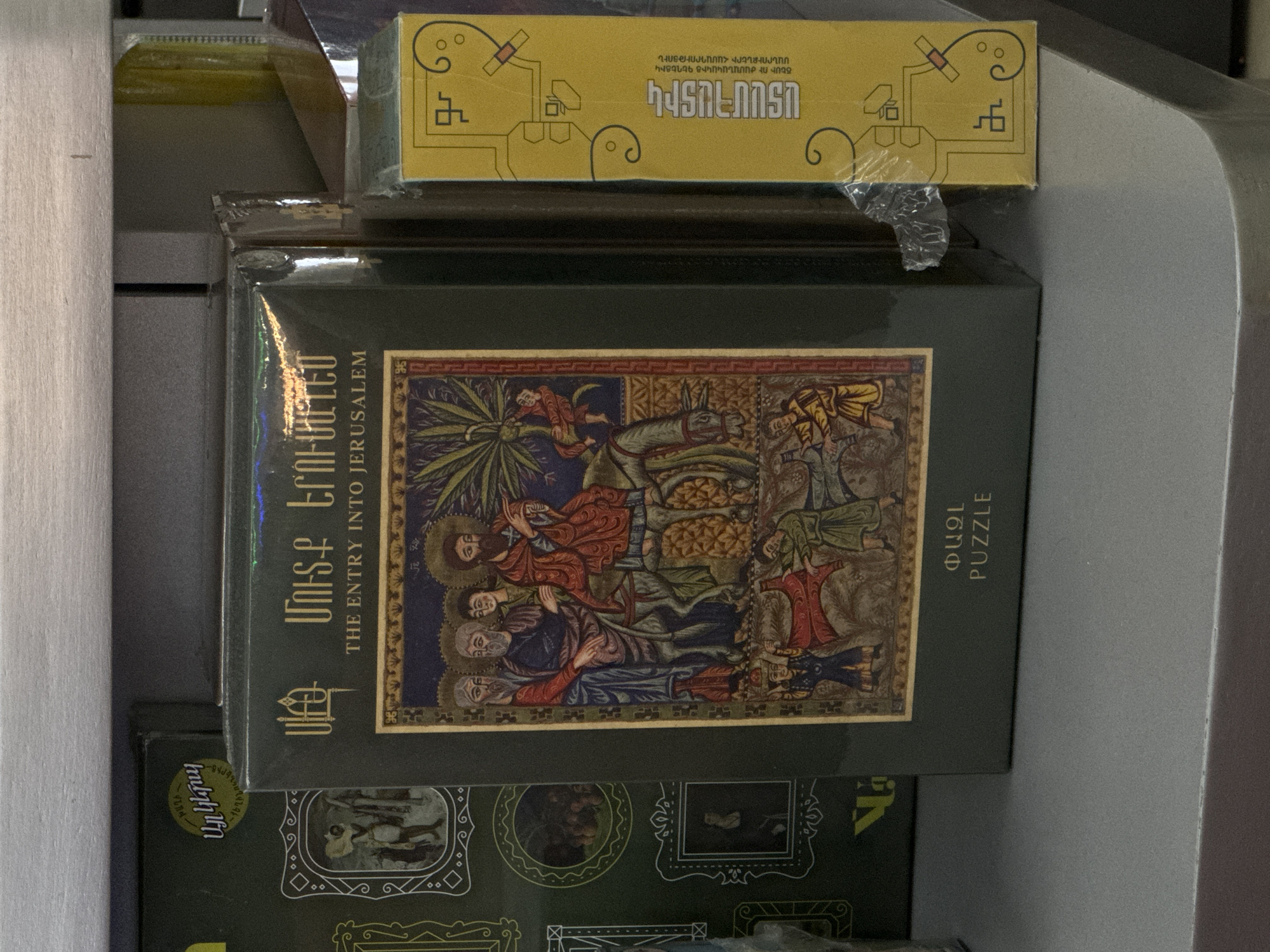 This image captures a stack of boxes, likely puzzles or games, displayed in an indoor setting. The central and most prominent object is a dark green rectangular box, oriented vertically, featuring an intricate illustration. The artwork depicts a biblical scene, specifically "The Entry into Jerusalem," with several haloed figures in ancient attire, one riding a donkey, and others laying cloaks or palm branches on the ground. The style is reminiscent of illuminated manuscripts or Byzantine art, characterized by rich colors like reds, blues, greens, and gold, and detailed patterns.
Above this main box, a smaller, yellow rectangular box is placed. It features abstract, geometric patterns in dark blue and several lines of Armenian text. Below the main box, parts of other boxes are visible, including one with a circular green label bearing the number "961" and the Armenian text "Այլ կերպ." A wooden vertical element is visible on the far left, suggesting a shelf or display unit.
Visible text on the main green box includes "ՄՈՒՏՔ ԵՐՈՒՍԱՂԵՄ" (Mutk' Yerusaghem), translated as "THE ENTRY INTO JERUSALEM," and "ՓԱԶԼ" (P'azl), translated as "PUZZLE." On the yellow box, the text includes "ԽԱՂԻ," "ԱՆԱԿԱՆԱԿԻՑ ԳԵՂԵՑԻԿ ՄԱՆՐՈՒՔՆԵՐԻ ՀԱՄԱԴՐՈՒԹՅՈՒՆ" (Unexpectedly beautiful combination of miniatures), "ԾԵՍԻ ՈՒ ԺԱՄԱՆԱԿԻ" (Of ritual and time/occasion), and "ՏԱՄՀԱՏԻԿ," likely a brand or product name. The overall scene suggests a retail environment where cultural or religious-themed products are sold.