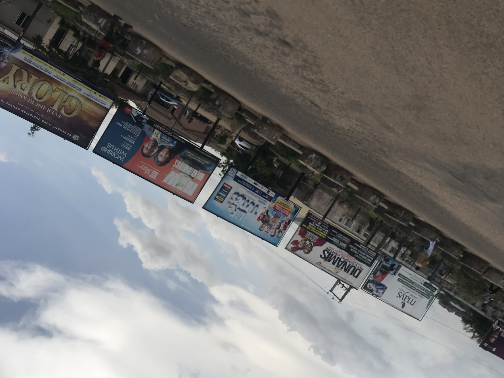 The image depicts a street scene in Jos, Nigeria, characterized by a row of billboards advertising religious services and events. The billboards are prominently displayed along the side of a paved road, suggesting an urban environment. The sky is partially cloudy, with patches of blue visible.
The billboards feature various messages and imagery. One large billboard proclaims "GLORY EVER-INCREASING" and mentions "COVENANT WORD CHRISTIAN CENTRE IN..." The text on this billboard is primarily in gold and brown tones, set against a sunset-like background. Another billboard, in shades of red and blue, has the heading "WORSHIP WITH US" and lists service times and a contact number. This billboard also features images of people, presumably congregants.
Further down the row, a billboard with the title "DUNAMIS INTL GOSPEL CENTRE" is visible. This billboard includes text about service programs and a phone number. Adjacent to it is a billboard that appears to read "mays" with a logo, and further information below it. Another billboard further down, partially visible, seems to have images of people and text that is difficult to decipher.
The road surface appears to be asphalt. There are some concrete structures along the roadside, possibly barriers or decorative elements. A few vehicles are visible on the road and parked nearby, including a white car and a dark-colored vehicle. In the background, there are buildings that appear to be commercial or residential structures, and some vegetation.
There are a few individuals visible in the scene. One person in a red jacket is walking on the sidewalk. Another person is partially visible further down the street. The overall impression is a dynamic urban setting with a strong presence of religious advertising. The angle of the photograph suggests it was taken from a moving vehicle or at a slight tilt.
