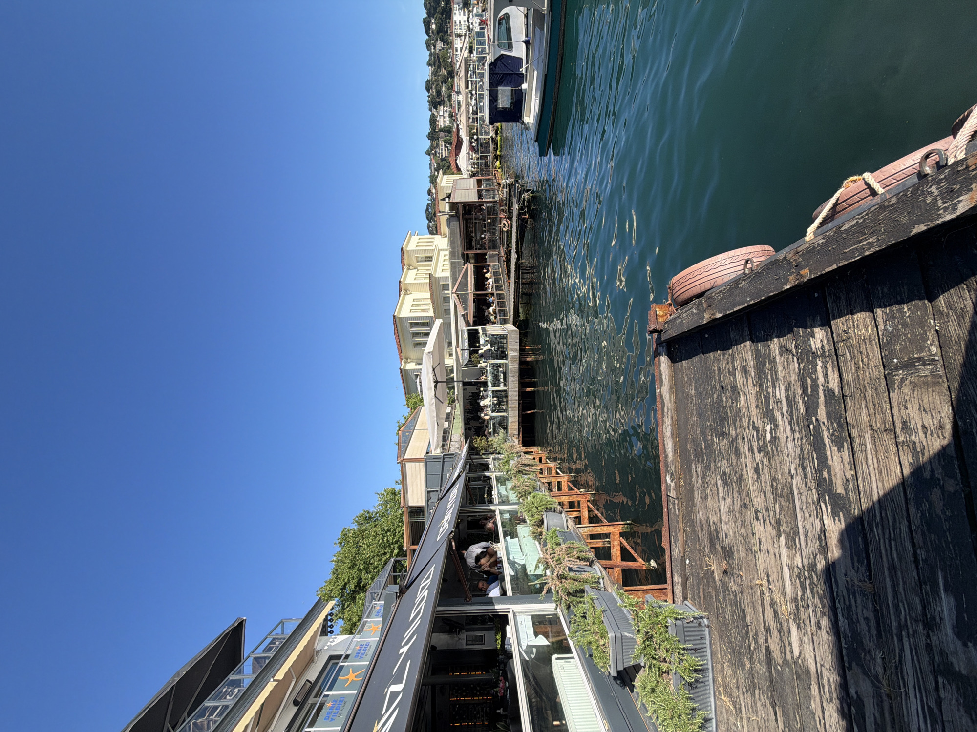 The image depicts a waterside dining establishment in Üsküdar, Turkey, likely on the Bosphorus strait, given the characteristic Ottoman-style architecture in the background. In the foreground, a weathered wooden pier with several planters containing green foliage leads towards the water. The pier is attached to a modern building with large windows, housing a restaurant called "DENİZ YILDIZI RESTAURANT," as indicated by signage. Several patrons are seated at tables both inside and on an outdoor deck area of the restaurant.
Adjacent to the restaurant, more dining spaces are visible, some with white umbrellas providing shade. The water is a deep teal color, with gentle ripples on its surface. A white and green boat is moored to the right side of the frame. Beyond the immediate structures, a hillside is dotted with residential buildings and trees. The sky is a clear, vibrant blue, suggesting a sunny day. The overall scene conveys a relaxed atmosphere of a waterfront cafe or restaurant.