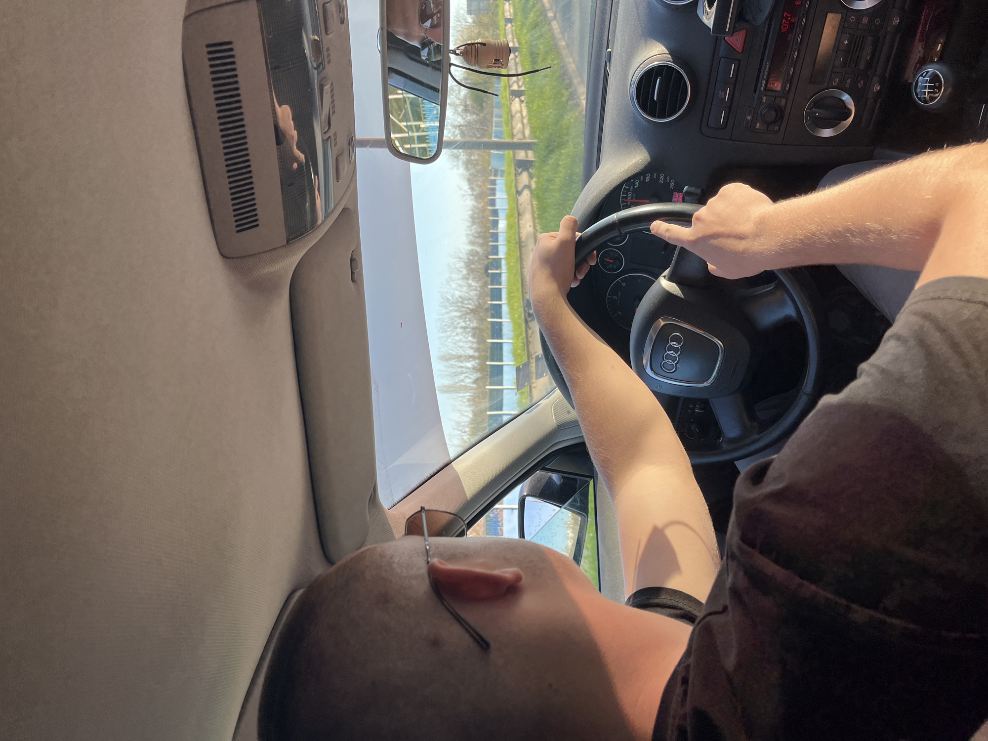 A man is driving an Audi. He is holding the steering wheel with both hands. He is wearing a black t-shirt and eyeglasses. The car is on a road with green grass on either side. The location is possibly Burgas, Bulgaria, based on the car's license plate and general environmental context. The dashboard is visible, showing a speedometer and a radio. A small air freshener is hanging from the rearview mirror.
