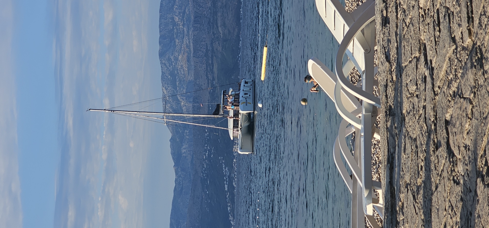 In the city of Grad Supetar, Croatia, on a sunny day with blue skies and scattered clouds, a catamaran is anchored in the calm blue sea. The catamaran, named "ANGELINA," has several people on its deck. In the foreground, two white lounge chairs are set up on a rocky shore, with a few people in the water, some appearing to be swimming. A yellow float is visible in the water near the catamaran, and a ball is floating nearby. In the background, a mountain range covered in vegetation forms the coastline. The image captures a serene coastal scene with recreational activities taking place.