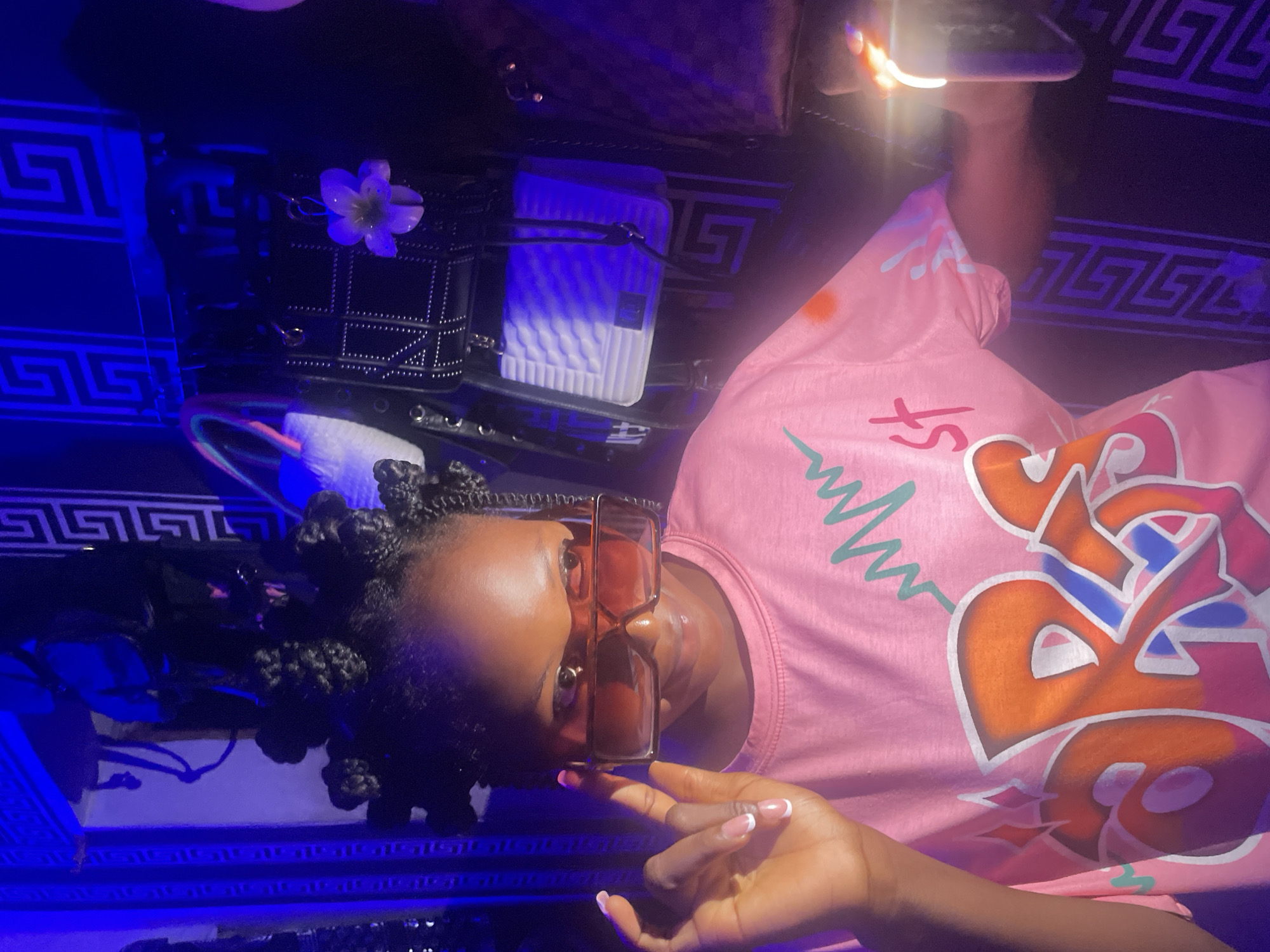 A woman with dark hair styled in small braids, wearing translucent orange glasses and a pink t-shirt with graffiti-style graphics, is positioned against a backdrop of a dark surface decorated with a Greek key pattern. The image has a blue tint. A black purse with a white flower attached and a white bag are also visible. The location is Lokoja, Nigeria.