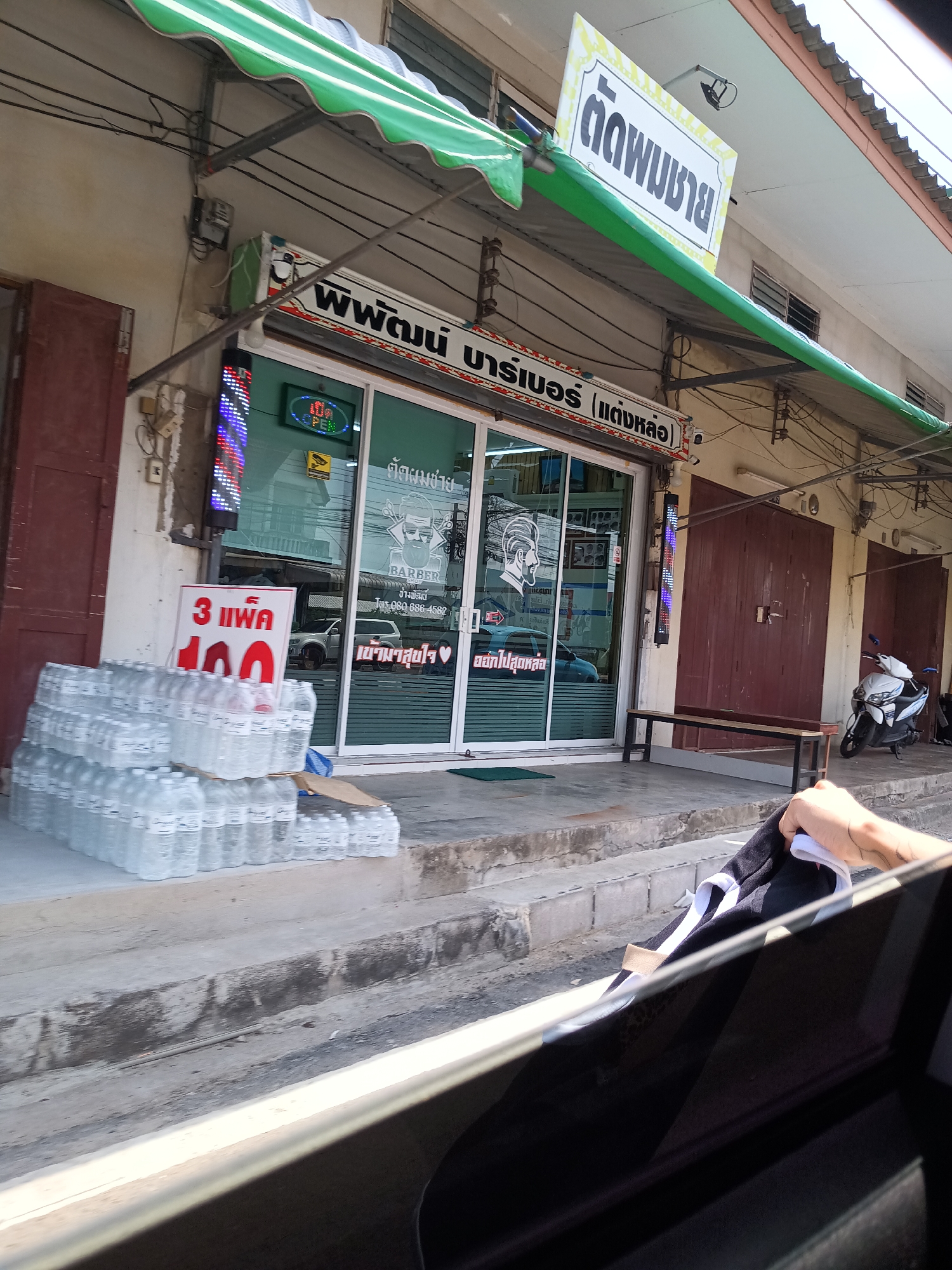 Here's a detailed description of the image:
**Location:** The image seems to be captured from inside a vehicle, looking towards a street in what is identifiable as a neighborhood in Hua Hin City Municipality, Thailand.
**Scene:** The main focus is on the front of a barbershop named "พิพัฒน์ บาร์เบอร์ (แต่งหล่อ)", which translates to "Pipat Barber (Handsome)". The shop has a green awning and large glass windows that display images related to barbering. There's a sign advertising a special offer for water bottles: "3 แพ็ค 100" (3 packs for 100). Stacks of bottled water are placed on the sidewalk in front of the shop.
**Objects:** A scooter is parked beside the barbershop. A bench sits beside the shop.
**People/Interactions:** No people are clearly visible, but a hand holding a piece of clothing is partially visible inside the vehicle in the foreground.