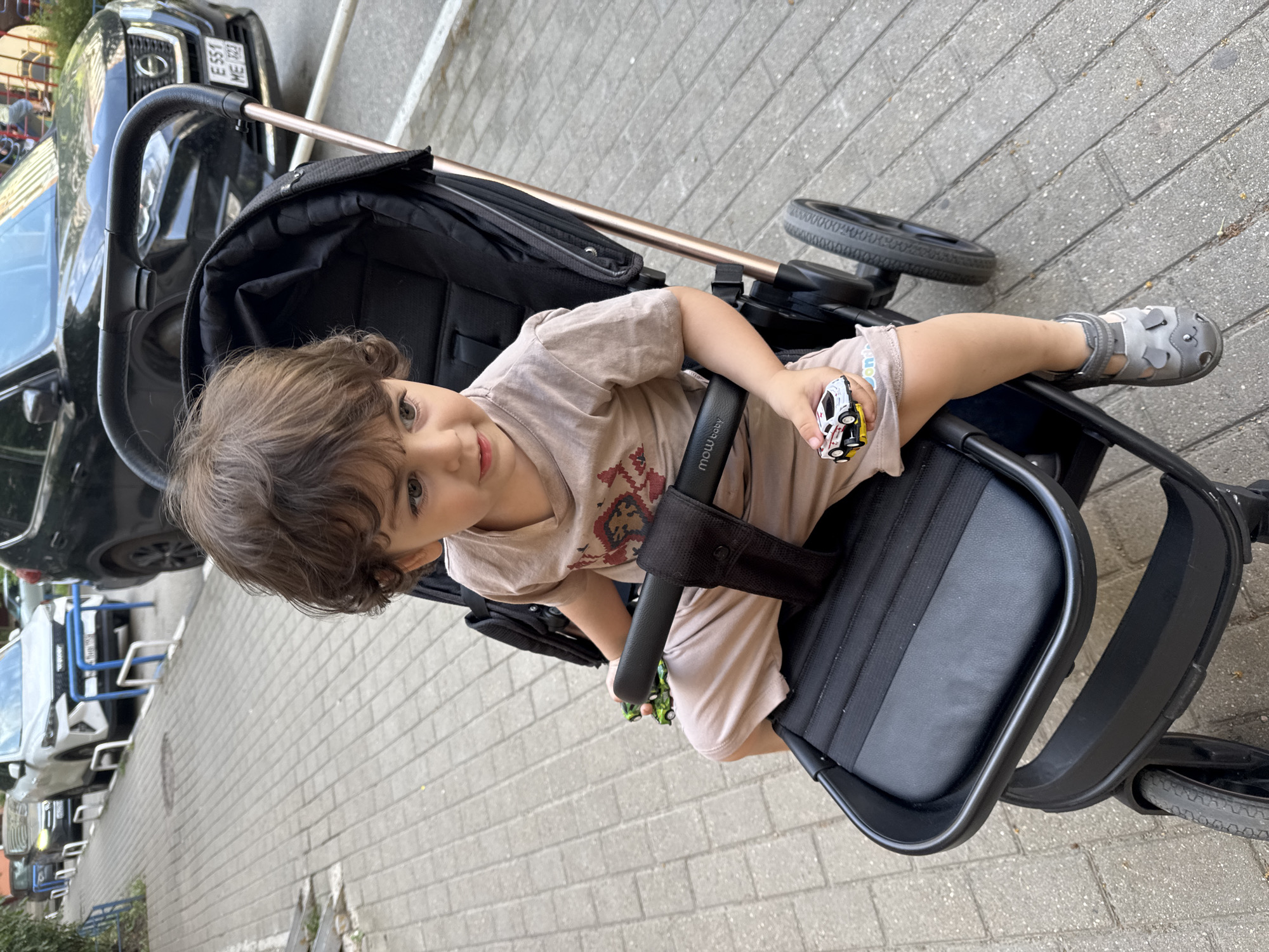 A young child with curly brown hair sits in a stroller on a sidewalk in Krasnodar, Russia. The child is wearing a light brown t-shirt and shorts, and grey sandals. The child is holding a small toy car in their right hand, and another toy car is visible in the stroller's tray. In the background, several cars are parked along the street, and a black SUV is parked directly behind the stroller. The sidewalk is made of grey brick pavers, and a metal railing with blue bars is visible in the foreground. The scene appears to be a sunny day.