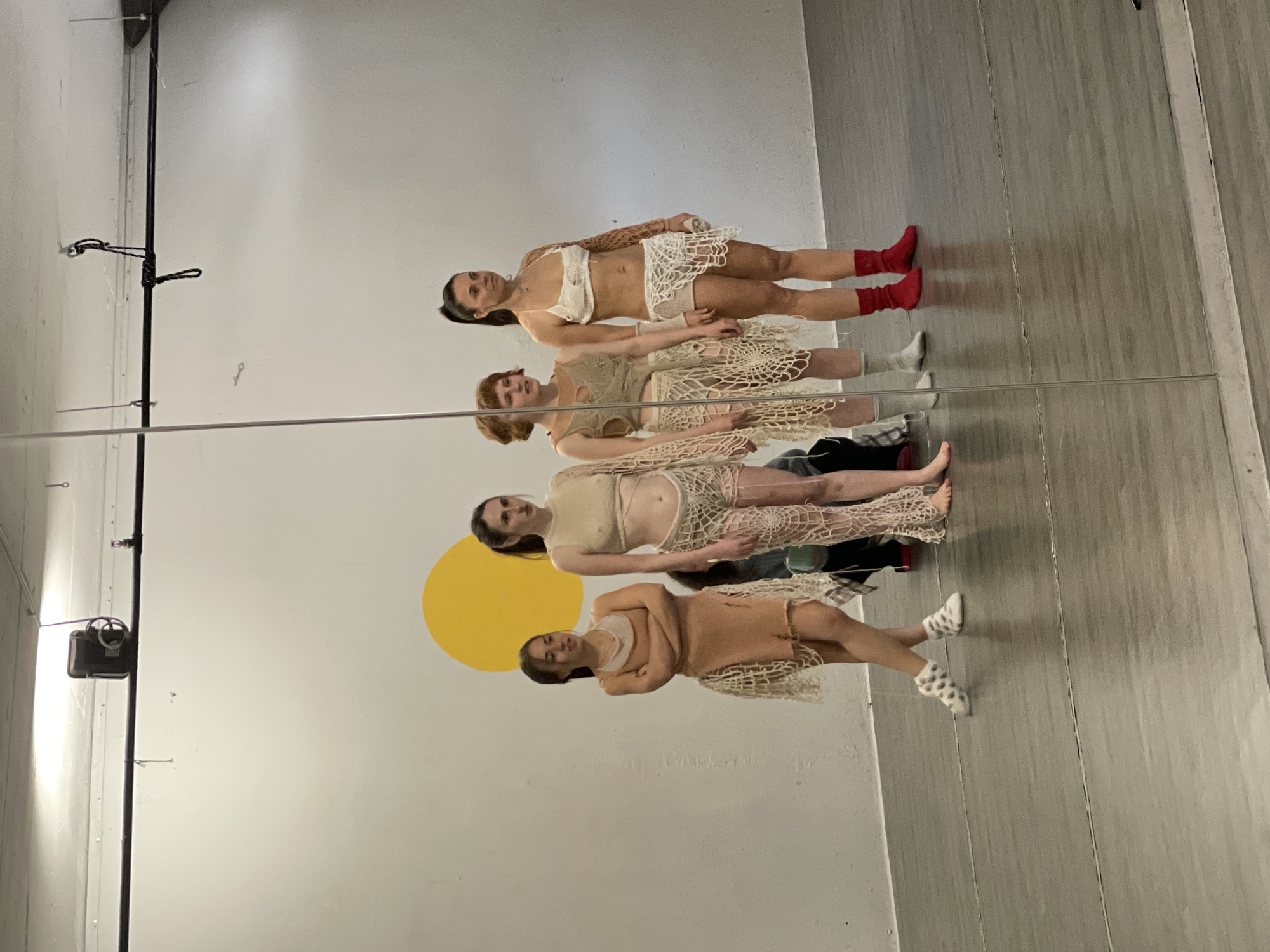 Four women stand in a line in front of a large mirror. All four women are wearing clothing that appears to be made of crocheted fabric. The woman at the top of the line is wearing a white top and skirt with red socks. The second woman is wearing a similar outfit that is tan in color. The third woman has on a tan top with shorts. The fourth women is wearing an orange-ish top and skirt with spotted socks. Behind the women is a white wall with a yellow circle painted on it. To the right is a wooden floor. The scene is indoors. The location is not discernible from the image provided.