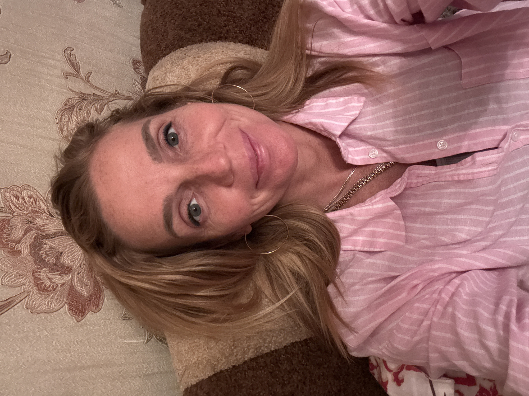 A woman with blonde hair and blue eyes, wearing a pink striped button-up shirt and gold hoop earrings, lies on a couch against a wall with floral wallpaper. She is looking directly at the camera and smiling. The image appears to be a selfie taken indoors, possibly in a residential setting. There is no visible indication of the specific location within Saint Petersburg, Russia.
