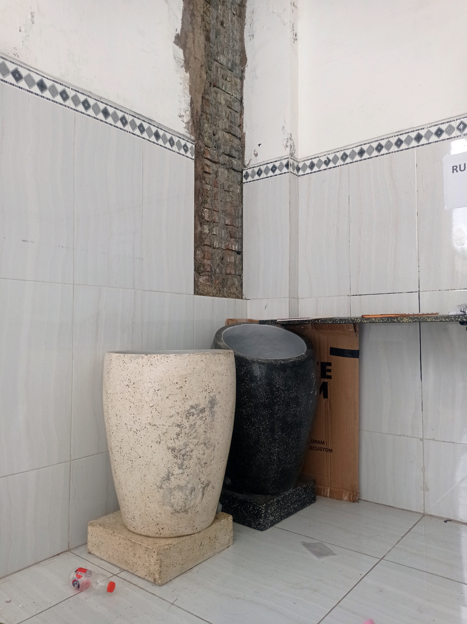 In Ganjaran, Indonesia, a corner of what appears to be an unfinished or undergoing renovation interior space is depicted. The walls are tiled with a light-colored, marble-patterned material, with a decorative border featuring a repeating diamond motif in grey and dark blue. A rough, exposed brick column rises vertically in the corner, showing signs of construction or disrepair.
In the foreground, two large decorative planters are positioned on the tiled floor. The planter on the left is a light beige, cylindrical vessel with a speckled texture. It rests on a square, beige stone base. To its right, a darker, black planter, also cylindrical with a speckled finish, is placed on a black stone base. The top opening of the black planter is visible and appears to be angled.
Behind the planters, a large piece of cardboard packaging stands upright, partially obscured by the black planter. Text is visible on the cardboard, including the word "GRAM" and "ECUSTOM." A narrow, speckled countertop extends horizontally along the back wall, above the cardboard.
On the tiled floor to the left of the beige planter, a discarded, clear plastic bottle with a red cap lies on its side. The floor itself is tiled with large, light grey square tiles. A sign with the text "RU" is visible on the right wall, partially obscured by glare. The lighting suggests an indoor environment, possibly with overhead or natural light sources not directly visible in the frame. The scene conveys a sense of temporary storage or an incomplete construction project.