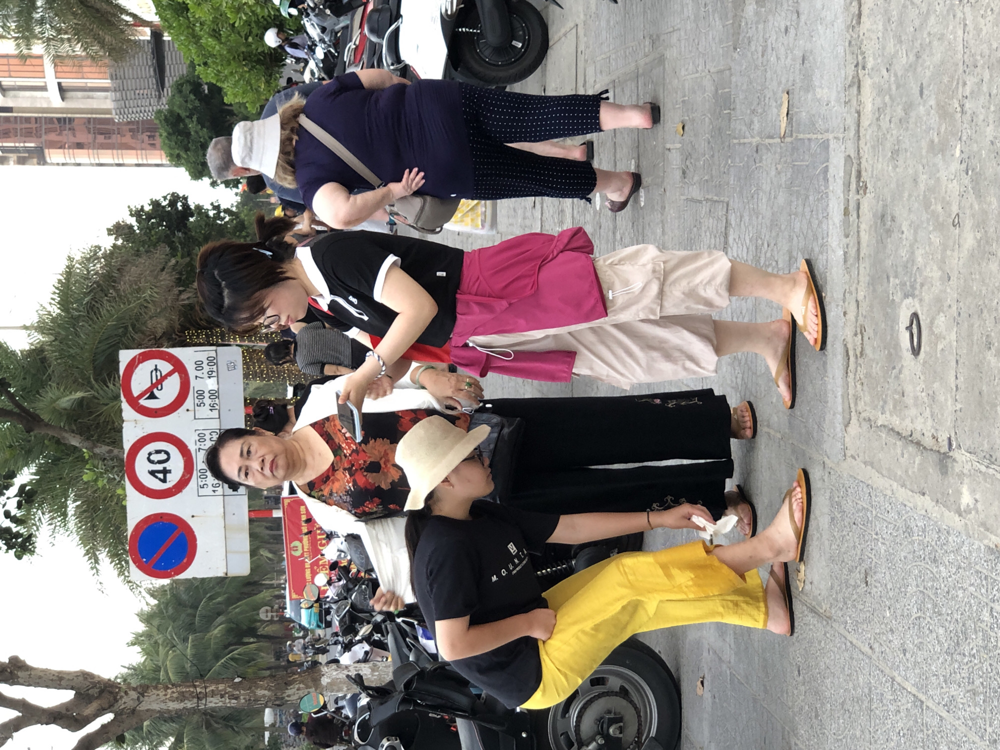 In Đà Nẵng, Vietnam, on a paved street with palm trees and buildings in the background, three women are the primary subjects. The woman on the left, wearing a black t-shirt and bright yellow pants, is bent over and appears to be picking something up from the ground. She has a straw hat on. The woman in the middle, wearing a white jacket over a floral print top and black pants, stands with her arms behind her back, holding a smartphone in her left hand. The woman on the right, wearing a dark blue t-shirt and patterned capri pants, has her left hand on her hip and is looking away from the camera. She has a white bucket hat on. In the background, a traffic sign displays "40" and a no-honking symbol, indicating speed limits and traffic regulations. Several motorcycles are parked to the left of the frame. The ground is made of grey paving stones, and there are some fallen leaves scattered about. The lighting suggests it is daytime, possibly late afternoon.