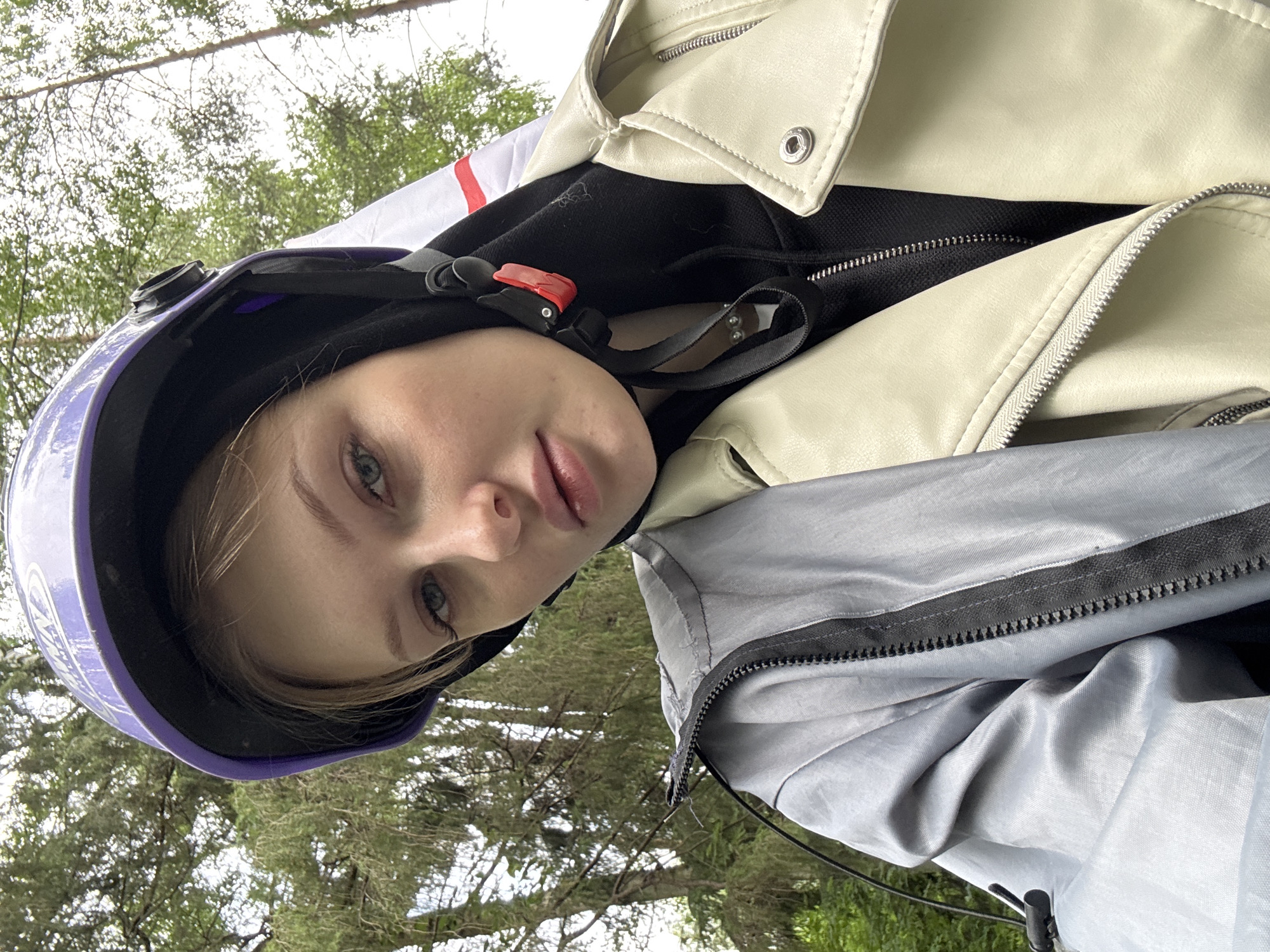 A young woman is depicted in a close-up, upward-angled selfie, set against a backdrop of dense forest foliage. The exact location in Perm, Russia, cannot be visually confirmed from the image itself.
The woman has light skin, light-colored eyes, and light blonde or brown hair tucked beneath a purple and black half-shell helmet. Her full lips are slightly parted, revealing a neutral expression as she gazes directly at the camera. She wears a black balaclava or hood beneath the helmet, and the helmet's black straps converge with a prominent red buckle visible on the viewer's left side of her chin. A light-colored, multi-beaded necklace is visible below the chin strap.
Her attire consists of layered jackets. The innermost visible layer is a black zippered garment, likely a hoodie. Over this, on her left side, she wears a light grey or silver jacket with a visible black zipper and a drawstring cord. On her right side, an outer jacket of cream or beige faux leather in a biker style is visible, featuring a prominent lapel, a silver snap button, and a zipper detail.
The background consists of numerous green trees, suggesting a forest or heavily wooded area under what appears to be an overcast sky.