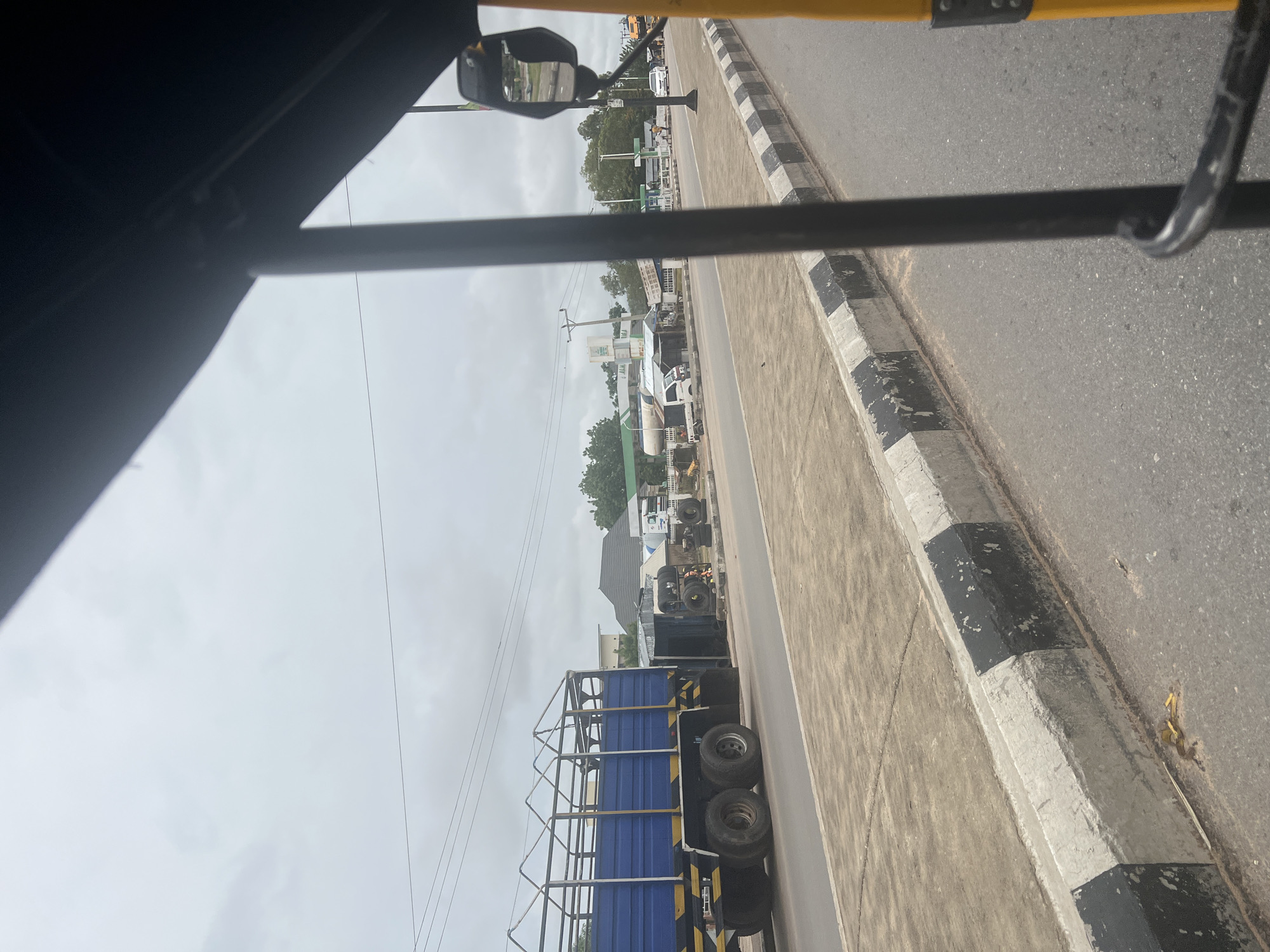 The image shows a view from inside a three-wheeled vehicle, likely a tuk-tuk, looking out at a street scene in Jalingo, Nigeria. The foreground is dominated by the yellow frame and black bar of the vehicle.
The street is paved with asphalt and has a concrete curb painted with alternating black and white stripes. On the side of the street, there is a blue truck with a metal cage-like structure on its back. Next to the truck, there are several stacks of tires and some smaller vehicles, including a white pickup truck and what appears to be a fuel tanker. In the background, buildings are visible, including what looks like a gas station with pumps and a small shop. Trees and utility poles line the street. The sky is overcast. A few people are visible in the distance, walking near the gas station. The overall scene suggests a commercial area on a cloudy day in Jalingo.