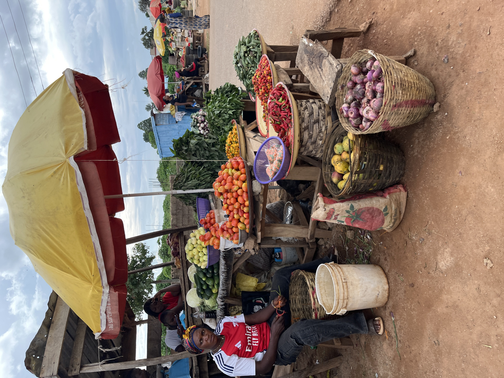 This image captures a bustling outdoor market scene in Jos, Nigeria. The foreground features a woman of dark complexion, seated on a low wooden stool to the left. She wears a red and white short-sleeved shirt with "Emirates Fly Better" branding, dark trousers, and open-toed sandals, topped with a yellow, blue, and red patterned headwrap. She holds a small, indistinct object in her right hand and looks directly towards the viewer with a neutral expression. Beside her are an empty woven basket and a white plastic bucket on the reddish-brown dirt ground.
Behind this woman, beneath a rustic wooden stall structure with a corrugated metal roof, another woman in a red shirt rests her head on her hand, leaning against a wooden counter. Another individual in a white shirt is partially visible behind her. Dominating the center of the image is a large yellow and red market umbrella, partially obscuring the stall's roof, with faint text reading "all starts with a..." on its fringe.
Beneath the umbrella and extending to the right, a vibrant display of fresh produce is arranged on wooden tables and stands. Piles of red and orange tomatoes are prominent in metal basins, alongside green cucumbers, white cabbages, and a mound of yellow-green fruits (possibly garden eggs). Large baskets on the ground contain purple-red onions and mangoes. Further to the right, other wooden stands hold bowls brimming with small red peppers, a pile of green okra, and substantial quantities of dark green leafy vegetables. A purple plastic basket on one table contains small, clear plastic bags with unidentified contents.
In the mid-ground, a blue corrugated metal container or shed is visible. Several people are scattered around this area, including a woman in a dark shirt standing near the blue container, and others under additional red and yellow market umbrellas further down a paved road. On the far right, a woman in a long, patterned blue and brown dress walks away from the main market activity. The overall setting is characterized by reddish-brown compacted dirt, a cloudy sky, and sparse distant trees. Power lines are visible overhead.