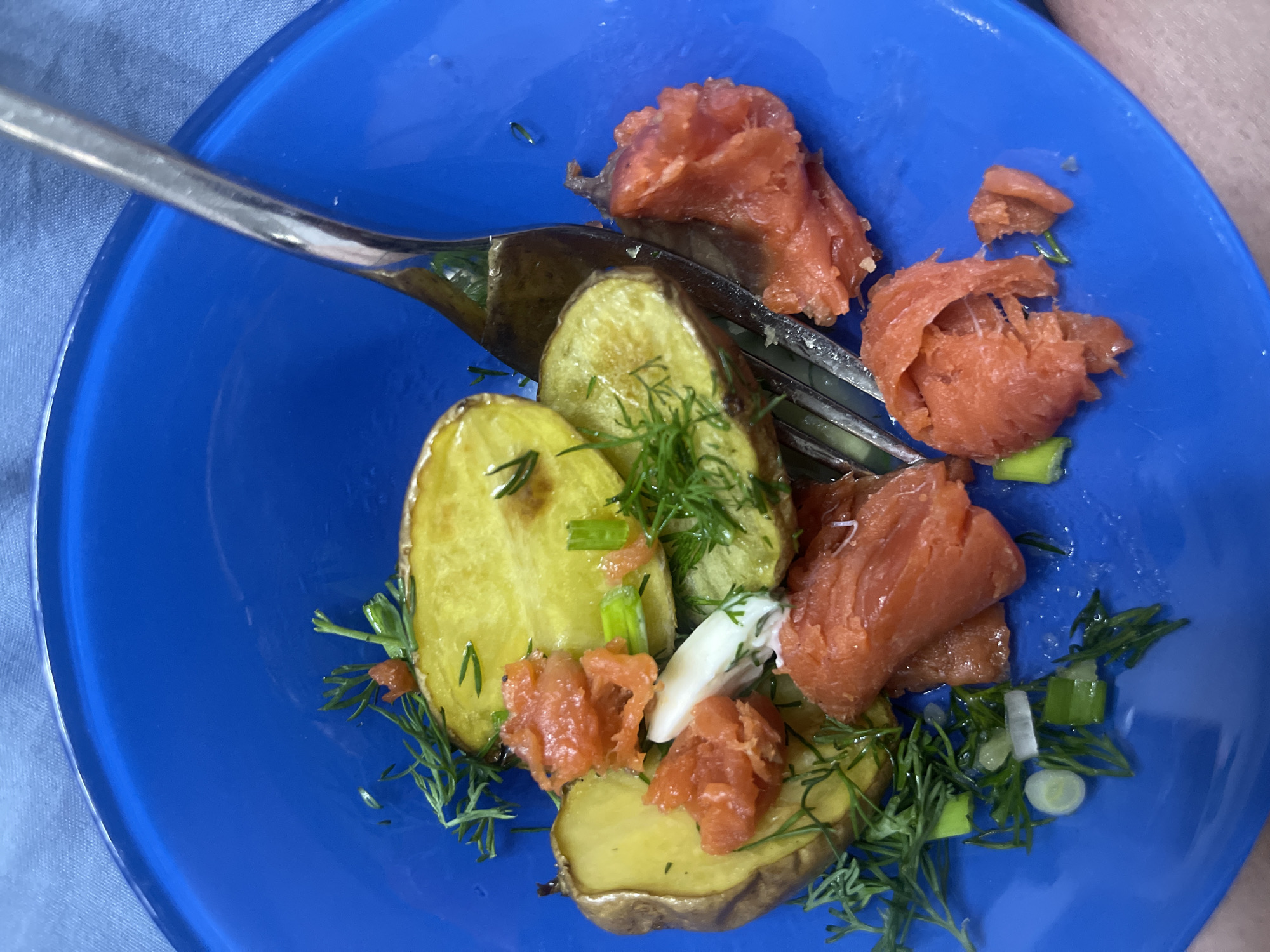 A close-up, overhead shot displays a prepared meal served on a round, deep blue plate. The meal consists of several sliced, skin-on potatoes, approximately golden-yellow in color, accompanied by numerous pieces of flaked, orange-red fish, consistent with salmon. Fresh green dill sprigs and chopped green onions are scattered over the food, along with a small dollop of a white, creamy substance. A silver-colored metal fork is positioned within the food on the plate. The plate rests on a light blue fabric surface, and a segment of a human leg or thigh is visible in the bottom right corner of the frame. The scene is located in the city of Chaikovsky, Russia.