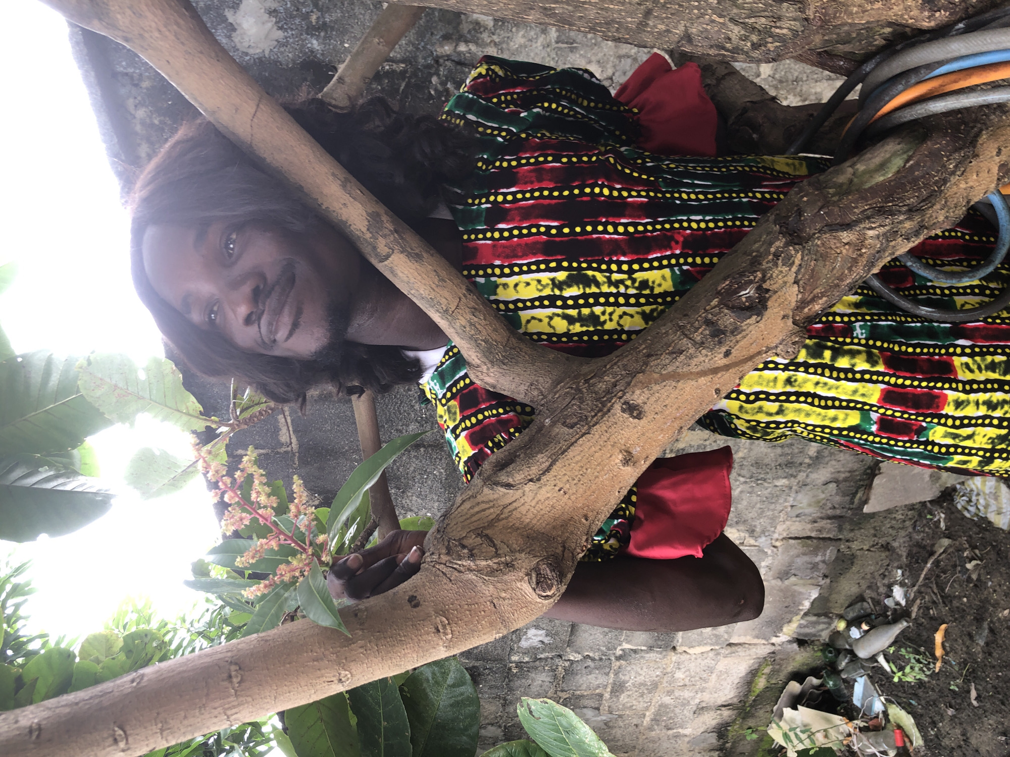 An African man with long, dark hair and a mustache stands outdoors in Ado, Nigeria, partially obscured by tree branches. He is looking upwards and to his left. He is wearing a colorful, patterned shirt with vertical stripes of yellow, red, and green, accented with small white dots. The shirt has red, ruffled cuffs. The background consists of a rough concrete wall and foliage, including leaves and a flowering branch held by the man's left hand. The ground below is dirt and appears to have some debris, including plastic bottles. Several cables or hoses are visible near the tree trunk on the right side of the frame.