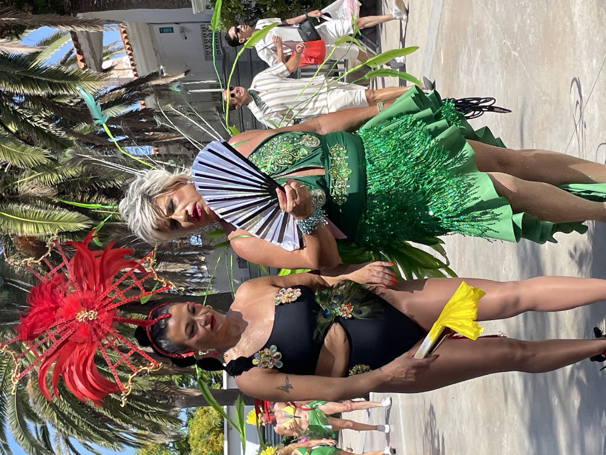 In Sitges, Spain, two women are dressed in elaborate costumes for a carnival parade. The woman on the left wears a black one-piece swimsuit adorned with floral appliques and peacock feathers, and a large, red feather headdress. She holds a yellow feathered fan in her right hand. The woman on the right wears a green sequined dress with a fringed skirt, elaborate green and gold embroidery on the bodice, and a silver-beaded bracelet. She holds a black and silver fan in her right hand and has a large feathered headdress with green and blue feathers. They are walking on a paved street lined with palm trees and buildings. In the background, two men in casual attire, one wearing a striped shirt and the other a white shirt, walk past. Other parade participants are visible in the distance.