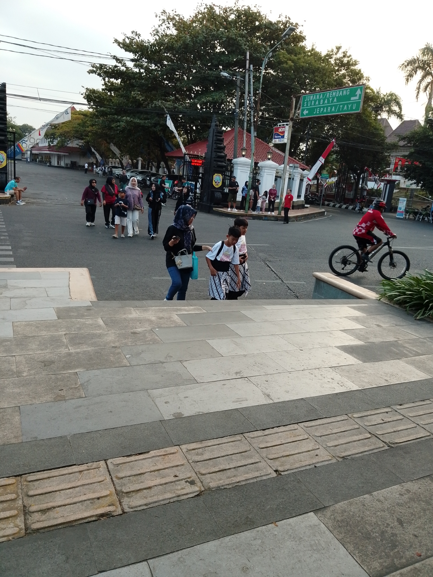 The image captures a street scene in Kecamatan Pati, Indonesia, characterized by an intersection with a traffic signpost and a group of pedestrians. The signpost displays destinations such as "JUWANA/REMBANG," "SURABAYA," and "JEPARA/TAYU," indicating it's a junction for travel. Several people are seen walking or standing near the intersection. In the foreground, a person is riding a bicycle in a red top and helmet. The environment includes trees, buildings, and a street, suggesting a public urban area. The perspective is from a slightly elevated viewpoint, possibly from a staircase, looking down towards the street. The paving stones on the steps in the foreground feature tactile paving for the visually impaired.