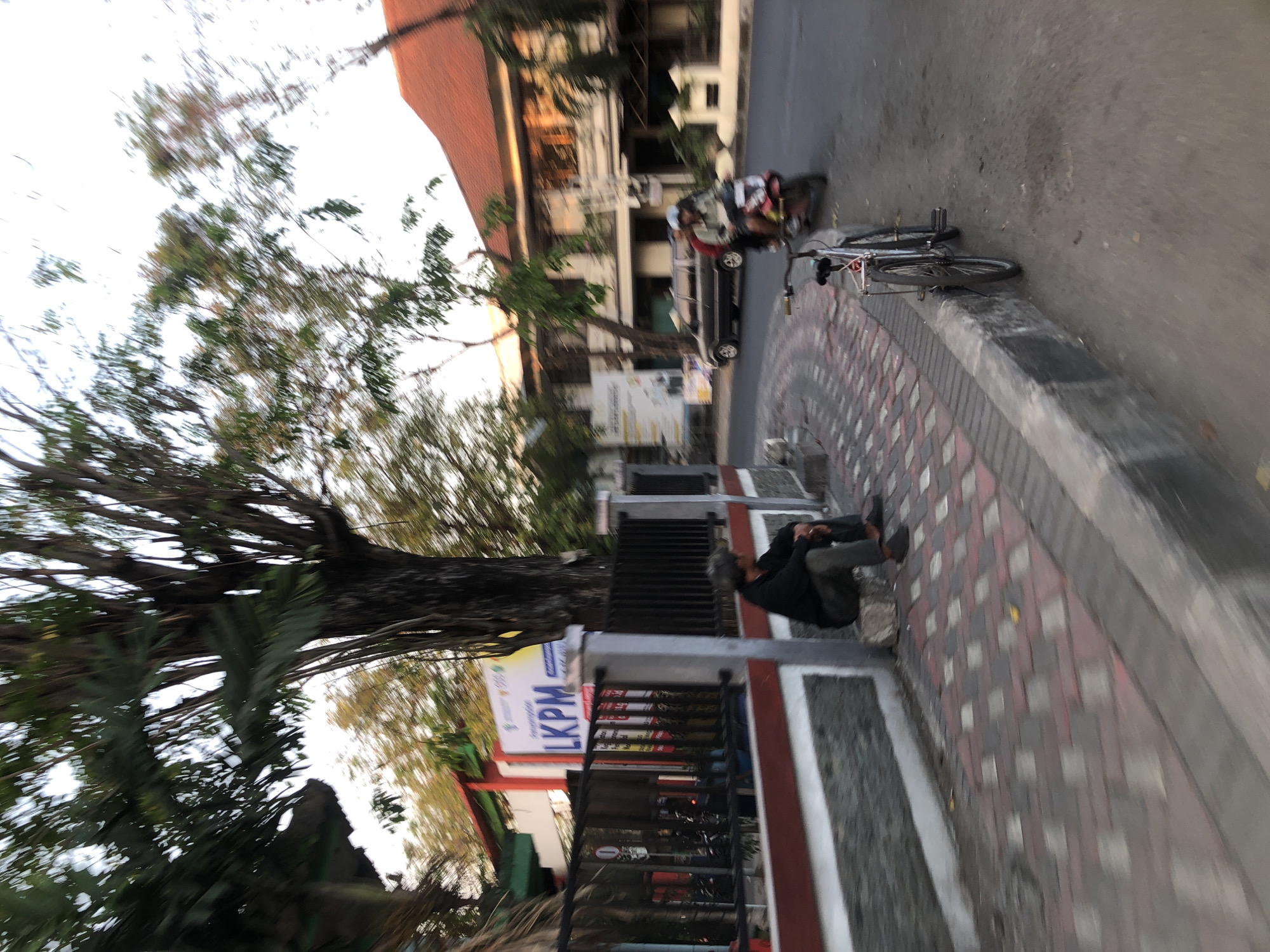 In Larangan, Indonesia, a person sits on a low wall beside a street. The individual, wearing dark clothing, is facing away from the camera and appears to be resting. The wall is constructed of stone and features a red and white border. The sidewalk is paved with red and gray interlocking bricks. Across the street, there is a building with a red tiled roof and white walls, possibly an office or residential structure. A dark-colored van is parked on the street, and a motorcycle with two riders passes by. Further down the street, a bicycle is parked next to the curb. Trees with dense foliage are visible along the street and near the buildings, suggesting a tropical or subtropical climate. The lighting indicates it might be late afternoon or early morning, with soft sunlight illuminating the scene.