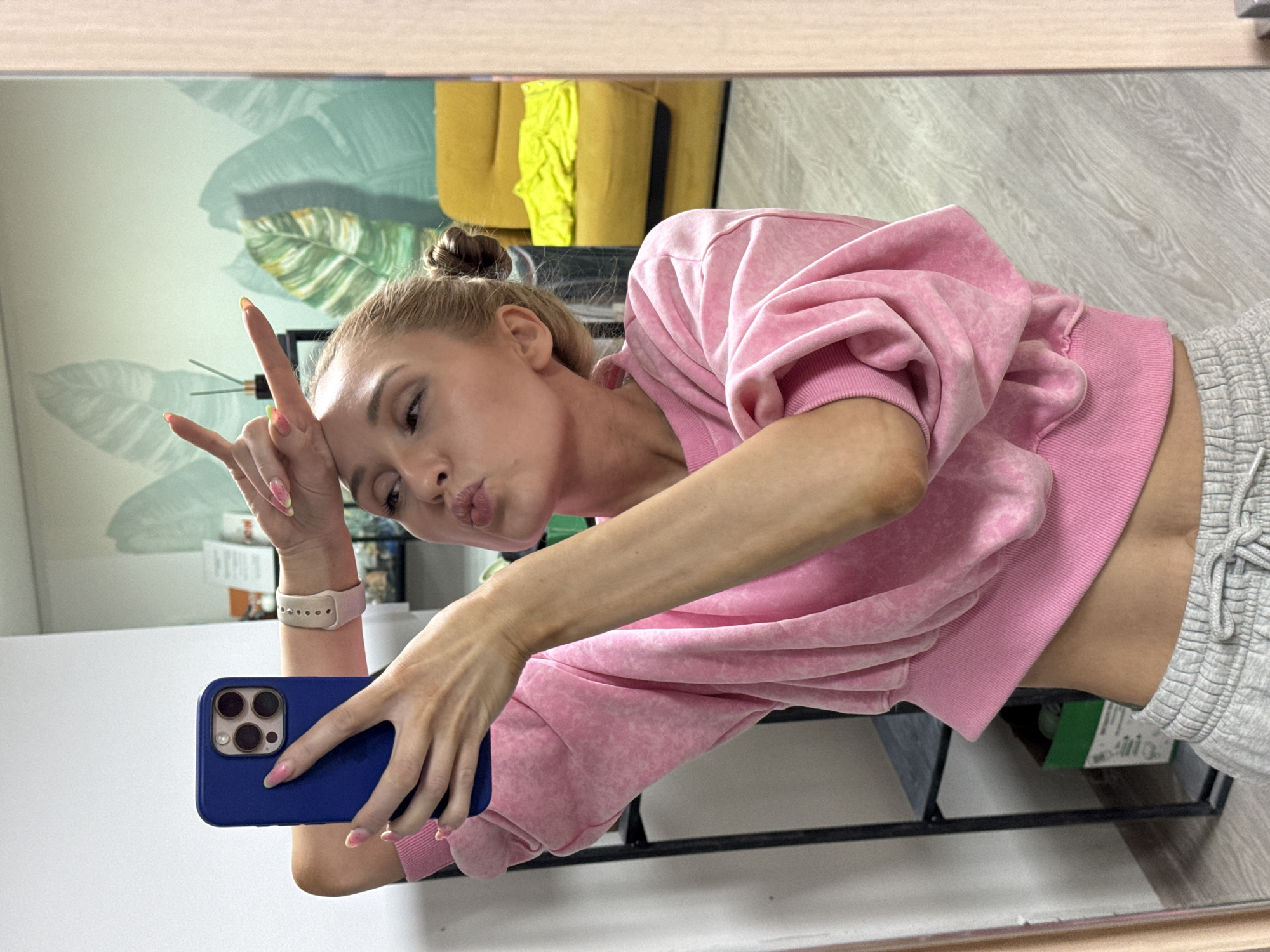 A young woman is captured in a mirror selfie, posing playfully in an indoor setting. She has light-colored hair styled in a bun and is wearing a pink, possibly tie-dyed or faded, cropped sweatshirt paired with light grey sweatpants featuring a drawstring. She holds a blue-cased smartphone, likely an iPhone due to its distinctive triple camera setup, in her left hand to take the photo. Her right hand is raised, forming a "rock on" or "devil horns" gesture, and she pouts her lips towards the camera. A light-colored smartwatch or fitness tracker is visible on her right wrist. Her fingernails are painted with colorful, possibly fruit-themed or abstract, tips.
The background, visible in the mirror's reflection, reveals a room with light green walls adorned with large, stylized leaf patterns, suggesting a natural or bohemian aesthetic. A mustard yellow armchair or couch is partially visible, along with light wood-patterned flooring. A reed diffuser with black reeds can be seen behind her head. The overall scene conveys a casual, self-expressive, and lighthearted mood. The setting appears to be a modern and comfortable living space, possibly a home in Chaikovsky, Russia. No visible text is present in the image.