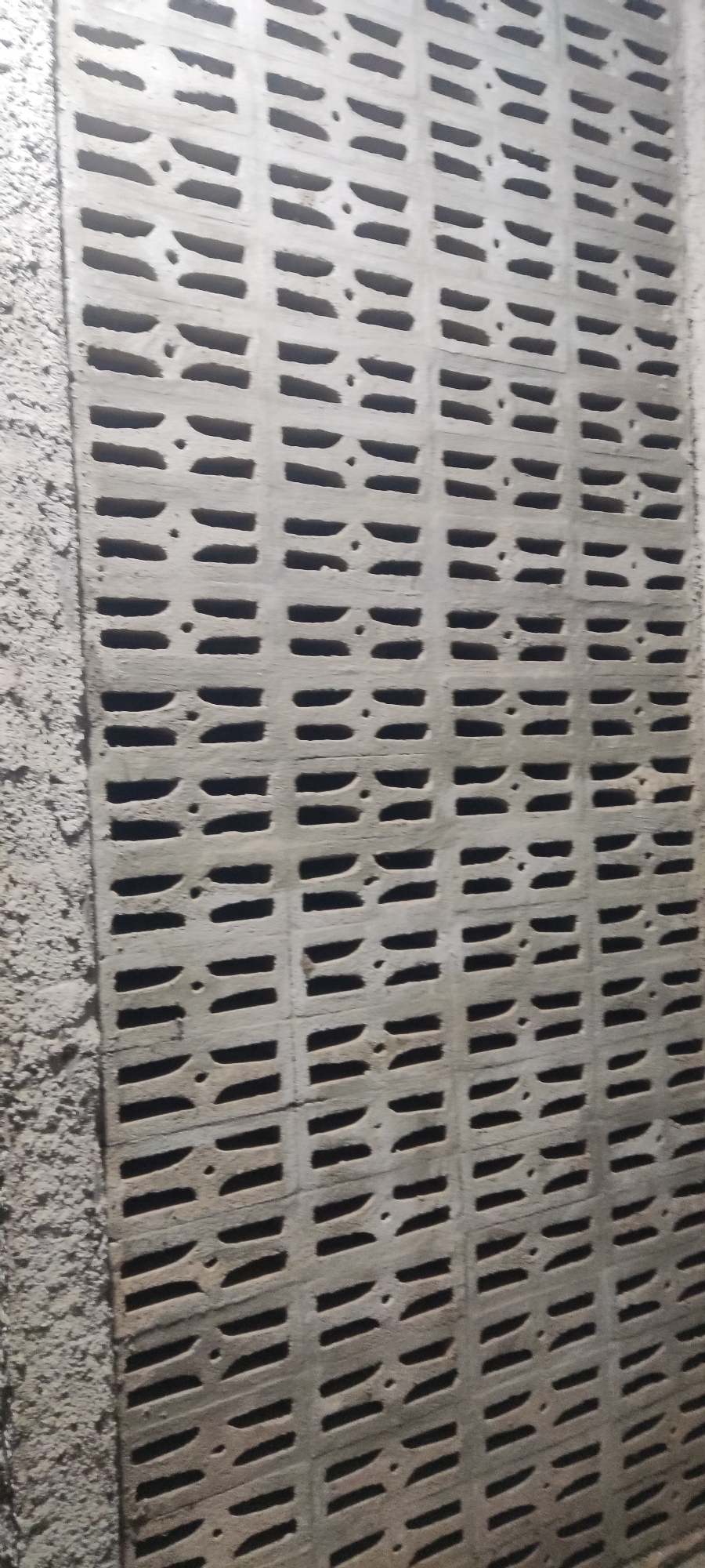 The image shows a close-up of a concrete block wall with a repetitive pattern of horizontal rectangular openings. The blocks are arranged in a grid, creating a lattice-like structure. To the left of the patterned wall, there is a rough, textured concrete surface. The lighting is dim, casting shadows within the openings and highlighting the texture of the concrete. No people or specific objects are visible, other than the wall itself. The scene appears to be outdoors or in a poorly lit indoor space, possibly a construction site or an exterior wall. The location of Karangwangun, Indonesia, is mentioned in the prompt but is not visually evident from the image itself.