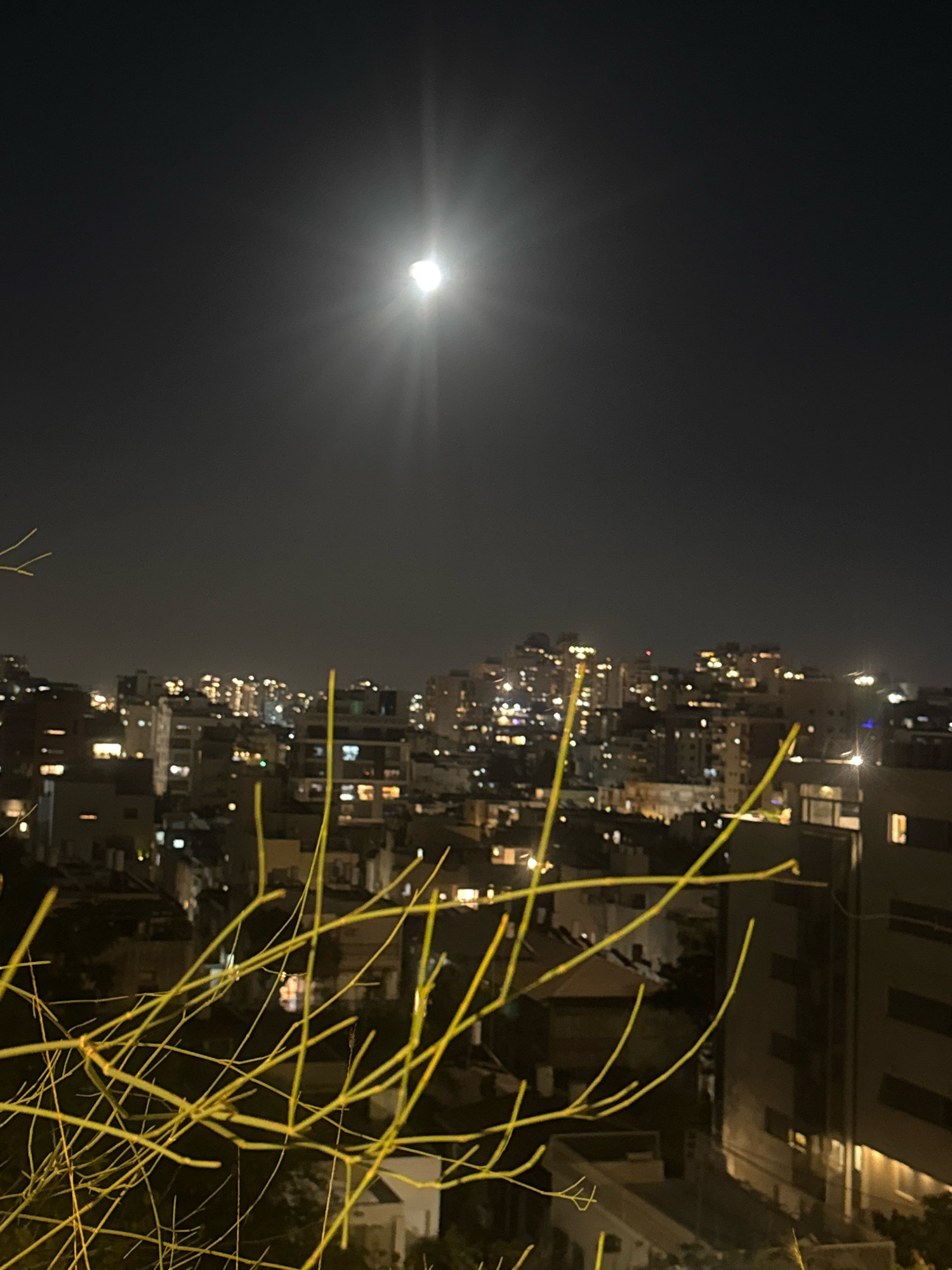 The image captures a night scene in the city of Ramat Gan, Israel, characterized by a bright full moon illuminating the sky above a cityscape. The moon is positioned high in the frame, emitting a strong glare that creates a starburst effect. Below the moon, the urban landscape unfolds with numerous buildings, many of which are lit from within, suggesting residential or commercial occupancy. The architecture appears to be modern, with varying building heights and styles. In the foreground, the skeletal branches of a tree, appearing almost neon yellow, crisscross the frame, creating a natural barrier and adding depth to the composition. The branches are bare, indicating a possible winter or arid season. The overall ambiance is one of quiet urban night, with the natural light of the moon dominating the scene. There are no people or discernible human activity in the frame. The environment is urban, with a dense concentration of buildings.