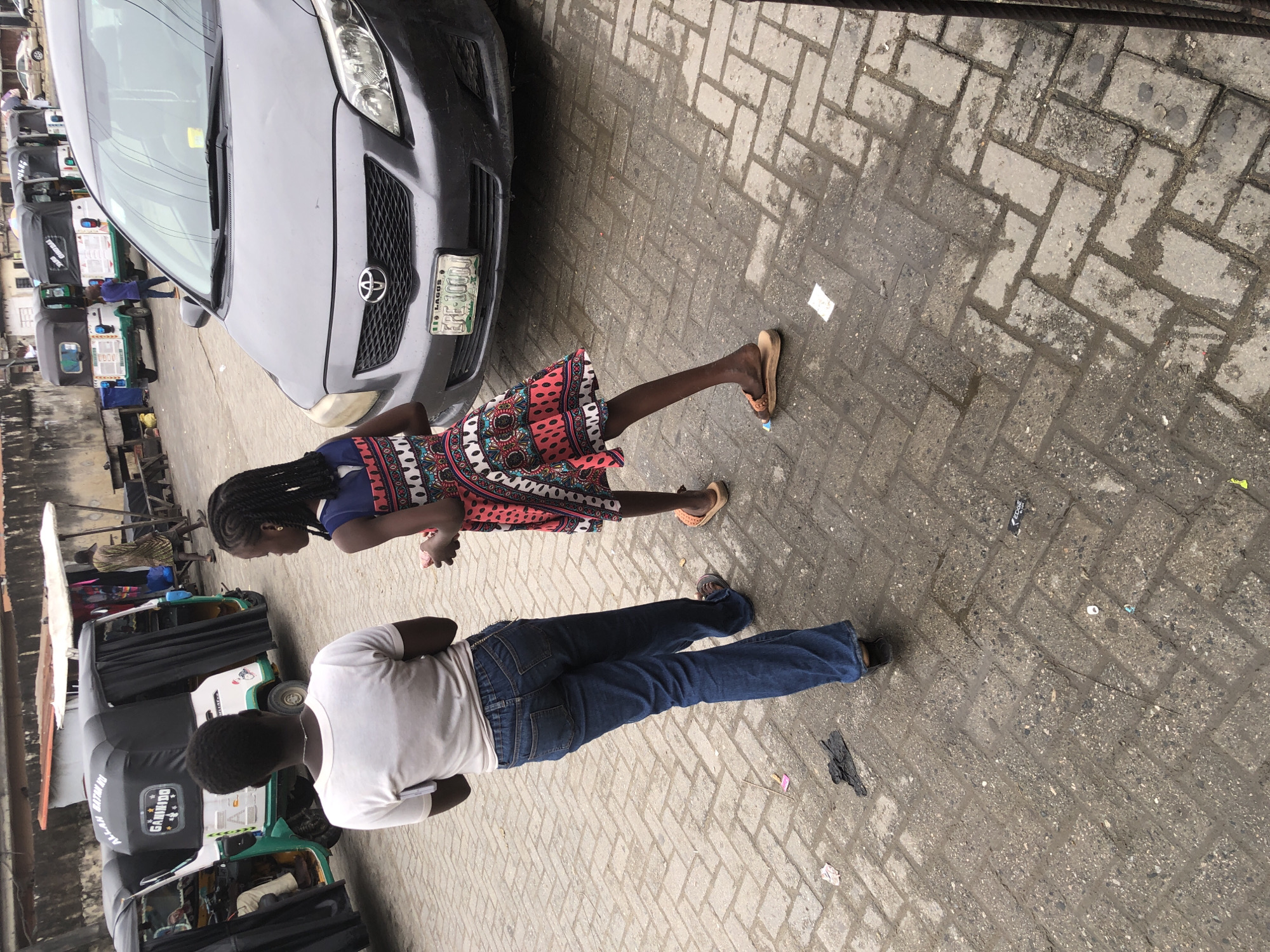 In the city of Ado, Nigeria, a paved street scene unfolds. Two young girls, viewed from behind, walk away from the camera. The older girl, wearing a white t-shirt and blue jeans, carries a white object tucked into her waistband. The younger girl, in a colorful patterned dress and brown sandals, holds something small and pink in her right hand. They are on a wet, brick-paved street.
In the background, several auto-rickshaws, commonly known as "Keke Napep" in Nigeria, are parked. One of these vehicles displays the text "ALLAH GANMA DEI" and "GAMIDDO". Beyond the auto-rickshaws, a gray sedan with a license plate reading "LAGOS EPE 1000D" is visible. The street is lined with buildings, and a vendor stall with white awnings is set up further down the road. Scattered on the wet pavement are pieces of trash, including a black glove and pink wrappers. The overall atmosphere suggests a busy, possibly humid urban environment.