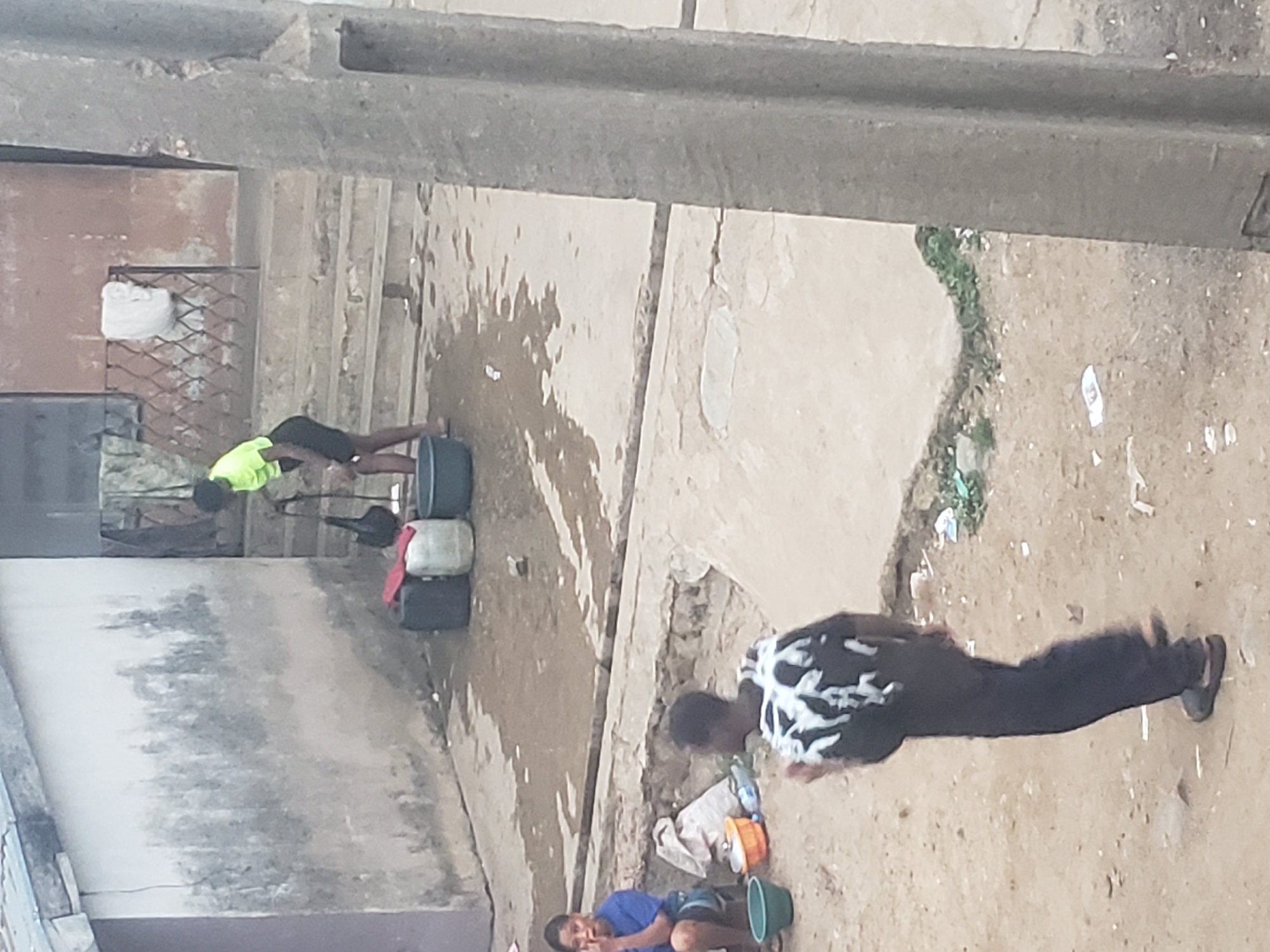 The image shows a street scene in Alalubosa, Nigeria. Three people are visible. One person wearing a yellow shirt is bending over containers. A second person in a black and white shirt is walking on the ground. A third person in a blue shirt is squatting by a bucket. The environment includes buildings, concrete, dirt, water, and various objects.