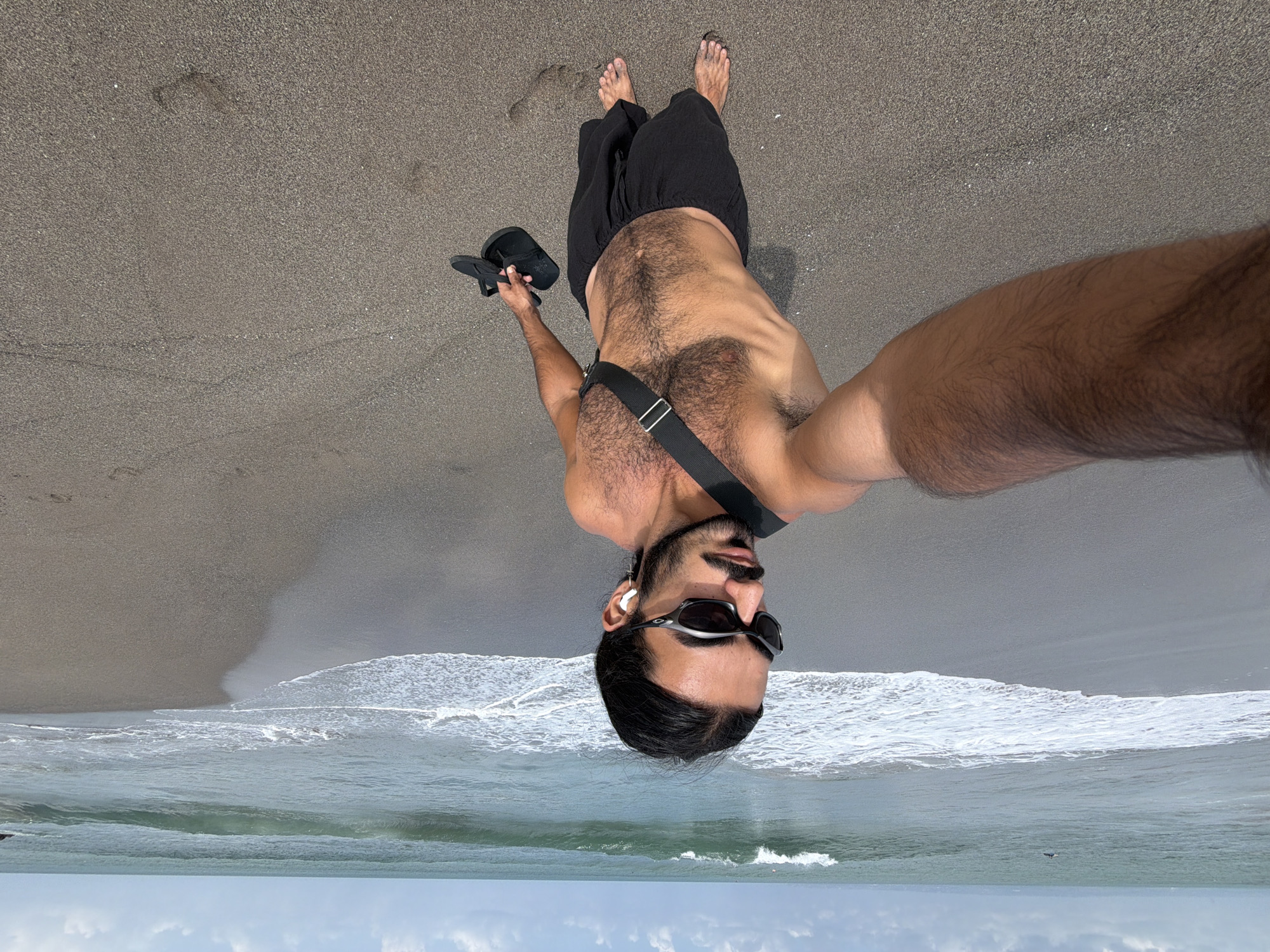 A man is taking a selfie on a beach in North Kuta, Indonesia. The man is wearing sunglasses, black shorts, and a black strap across his chest. He is holding a pair of black flip-flops in his right hand. The ocean is visible in the background with waves breaking on the shore. The beach is sandy and appears to be overcast.