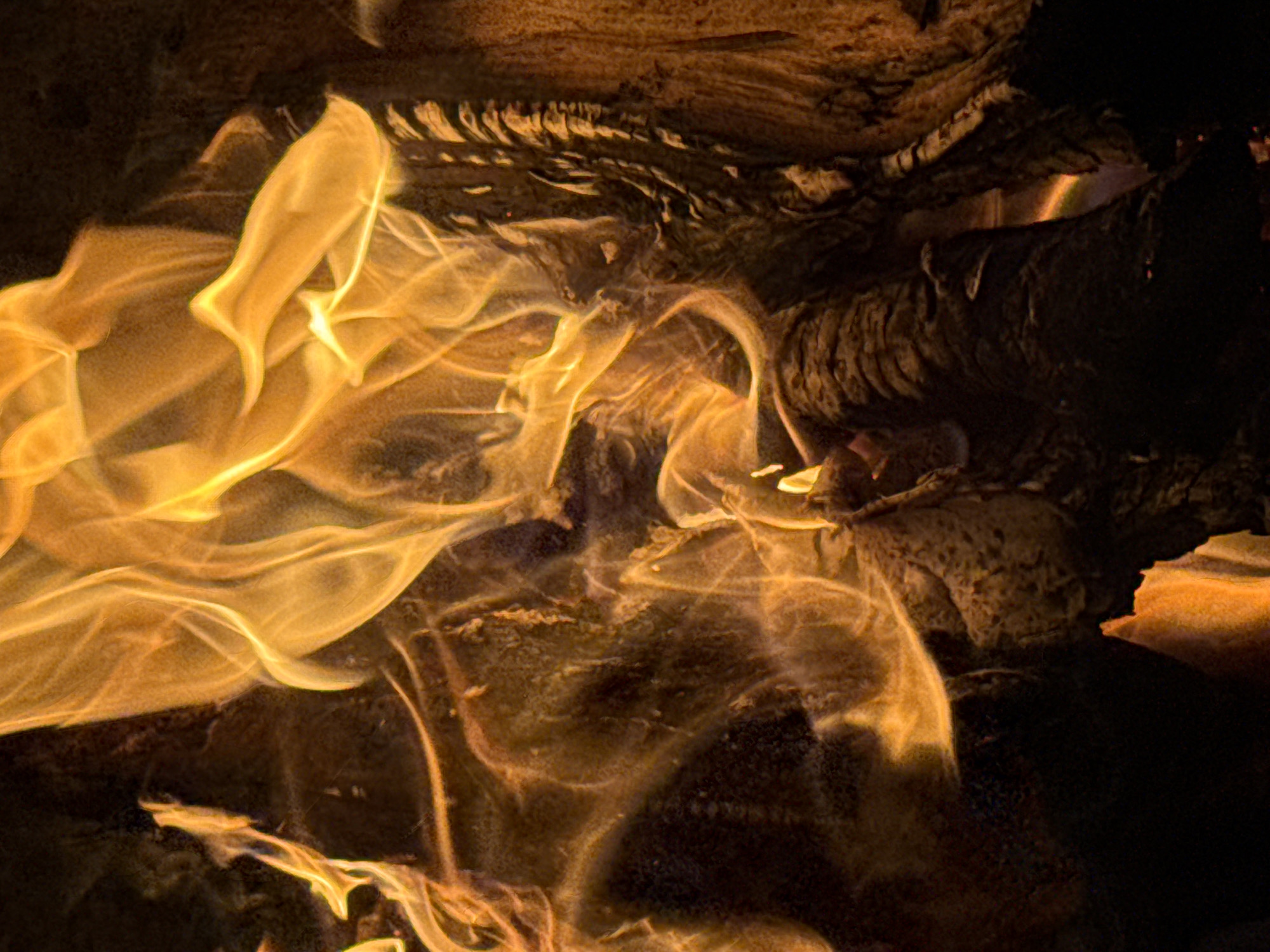 A close-up image depicts intense, stylized yellow and orange flames appearing to consume dark, coarse wooden logs or similar materials. The wood's surface is textured and cracked, with a combination of light and shadow highlighting its contours. The background fades to dark shades, suggesting the focus is on the burning material and the flames themselves. The scene suggests an uncontrolled burn, without any indication of any humans, and the location cannot be determined from the provided image.