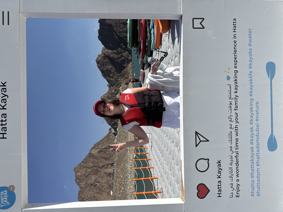 The image depicts an outdoor recreational scene at Hatta Kayak, located at the Hatta Dam in the city of Hatta, United Arab Emirates, characterized by a mountainous desert landscape and a large turquoise body of water.
In the foreground, a white, freestanding frame designed to resemble an Instagram post is prominently displayed. The top section of the frame features the text "Hatta Kayak," a circular blue logo with "HATTA KAYAK" and a plus sign, and a menu icon. The bottom section includes Instagram-style icons (heart, comment, share, bookmark), the text "Hatta Kayak," Arabic text, and English text reading "Enjoy a wonderful time with your family kayaking experience in Hatta," accompanied by two blue heart emojis. Below this, various hashtags related to Hatta and kayaking are listed, and at the very bottom, a graphic of two crossed light blue kayak paddles is present.
Centered within this frame is an adult female, likely posing for a photograph. She wears a red life vest with "KAYAK" text, a white t-shirt, white shorts, and a red baseball cap. Her left hand is raised, displaying a peace sign. Her right arm is bent, holding a white towel, a clear plastic water bottle, and black sunglasses. She stands on a gray, modular floating pontoon or dock, which extends into the water from the left side of the frame and features orange railings.
The background consists of a clear blue sky and rugged, brown rocky mountains, typical of the Hajar Mountains in Hatta. Below the mountains, the calm, turquoise water of the Hatta Dam is visible. On the floating platform to the right of the woman, a stack of colorful kayaks and small boats (green, red, orange, pink, light blue) is present. Further in the background, on the water, several indistinct individuals are observed on kayaks and paddleboards, engaged in water activities. The scene indicates a sunny day, suggesting clear weather conditions for outdoor recreation.