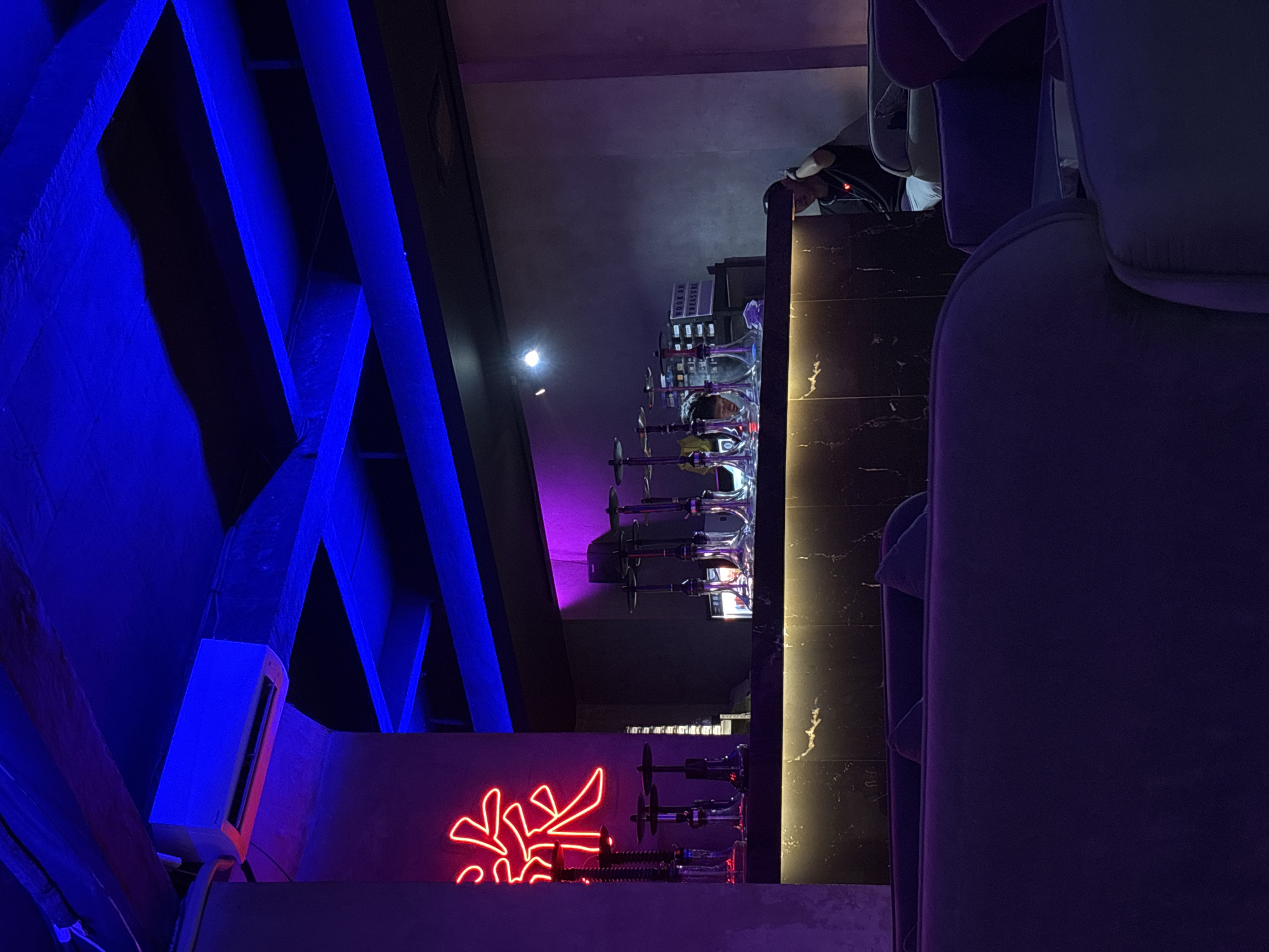 The image captures an indoor scene, likely a modern lounge or bar, characterized by dim lighting and vibrant accent colors. The perspective is slightly elevated, looking across a room.
On the left side of the frame, several prominent architectural beams are illuminated with bright blue LED strip lights, creating a striking visual effect against the dark background. An air conditioning unit is mounted on a wall structure beneath these beams. Below the air conditioner, a red neon sign displays a stylized character or symbol, possibly Asian script.
To the right of the blue-lit beams and neon sign, a long, dark counter stretches across the midground. Several hookahs (shisha pipes) are neatly arranged on this counter, each featuring clear glass bases and metallic stems, some appearing to have purple illumination. Behind the counter, a person is partially visible, standing and seemingly attending to the area, though their face is obscured. A small sign behind the hookahs reads "HOOKAH TREASURE."
The right side of the image features a seating area with plush, dark-colored couches or chairs, providing a comfortable and inviting atmosphere. A dark marbled wall or counter surface, illuminated by a warm, yellowish light strip, runs alongside the seating area. The overall ambiance is cool and sophisticated, with dominant hues of deep blue, purple, and black, suggesting an evening or nighttime setting. No specific activities are explicitly shown beyond the general setup for a lounge environment.