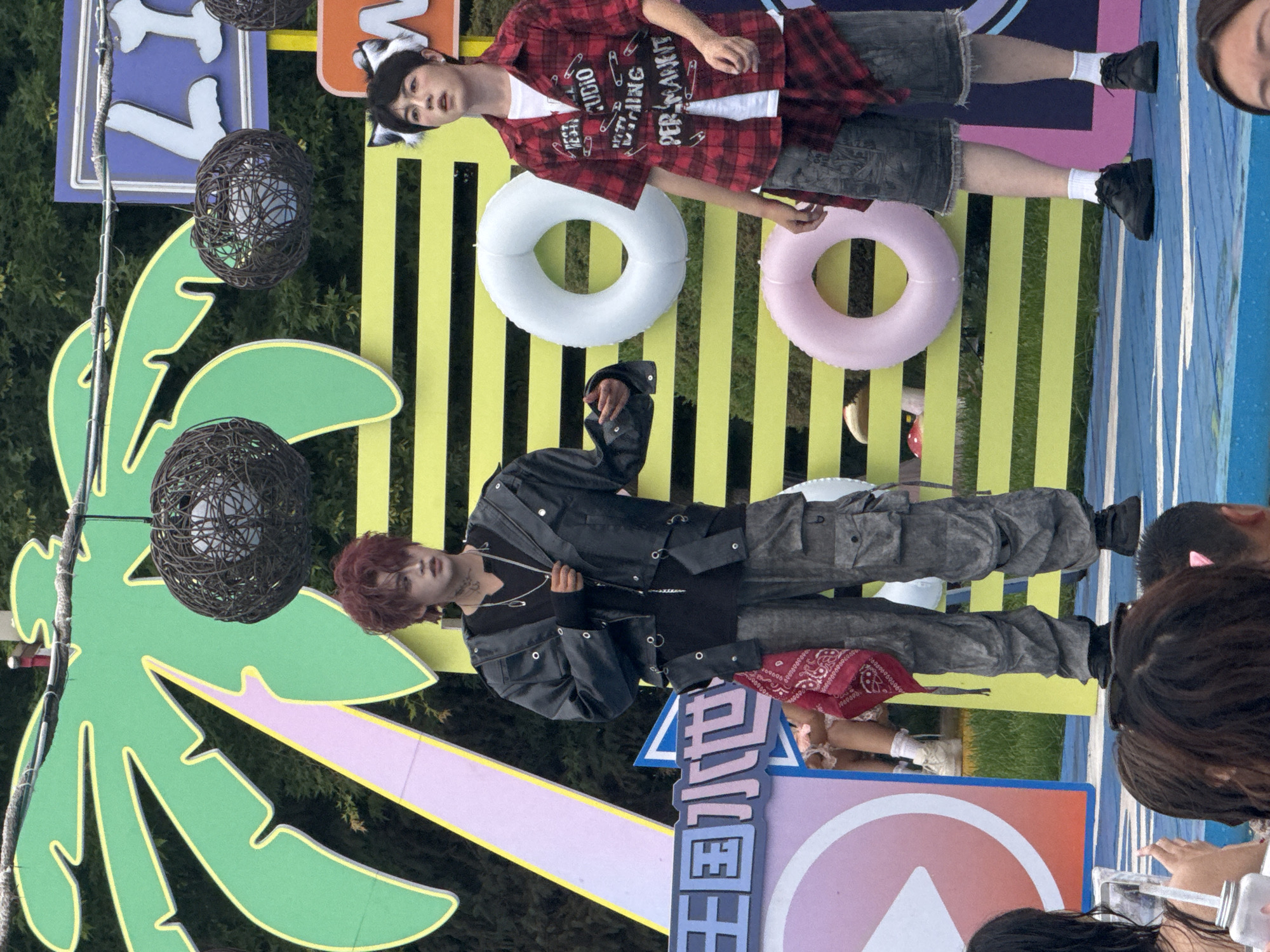 The image depicts an outdoor event, possibly a music performance or festival, set against a backdrop featuring a large green palm tree cutout with yellow and pink accents. Two young individuals are the primary subjects, standing on a blue stage. The person on the left has red dyed hair and is wearing a dark jacket over a black shirt, grey cargo pants, and black shoes. They are accessorized with a red bandana tied around their waist and several necklaces. The person on the right has black hair and is wearing a red and black plaid shirt over a white t-shirt, dark shorts, white socks, and black shoes. They also have cat-ear accessories on their head. In the background, there are two inflatable rings, one white and one pink, hanging on a yellow striped structure. A sign with blue and white lettering is visible in the upper right corner. The lower portion of the image shows the backs of several audience members, some holding up phones. The setting appears to be a public space, with elements suggesting a playful or entertainment-focused atmosphere. No specific indication of Taihe District, China, is present in the visual information provided.