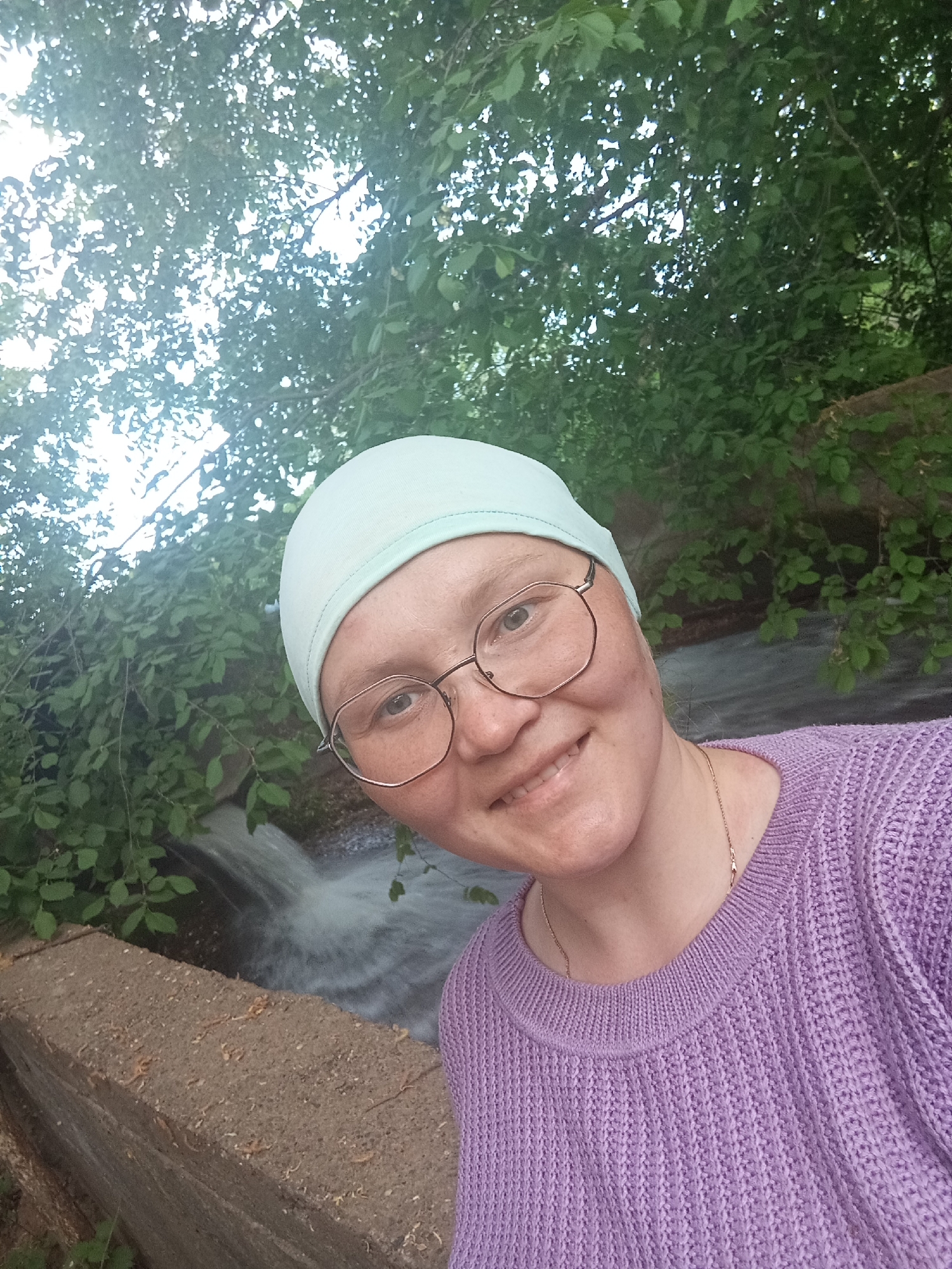 A person with glasses and a light blue head covering poses for a selfie. The person is wearing a lavender knit sweater and a delicate gold necklace. In the background, green trees overhang a stream of flowing water, partially obscured by a gray concrete barrier. The location is Unknown, Unknown.