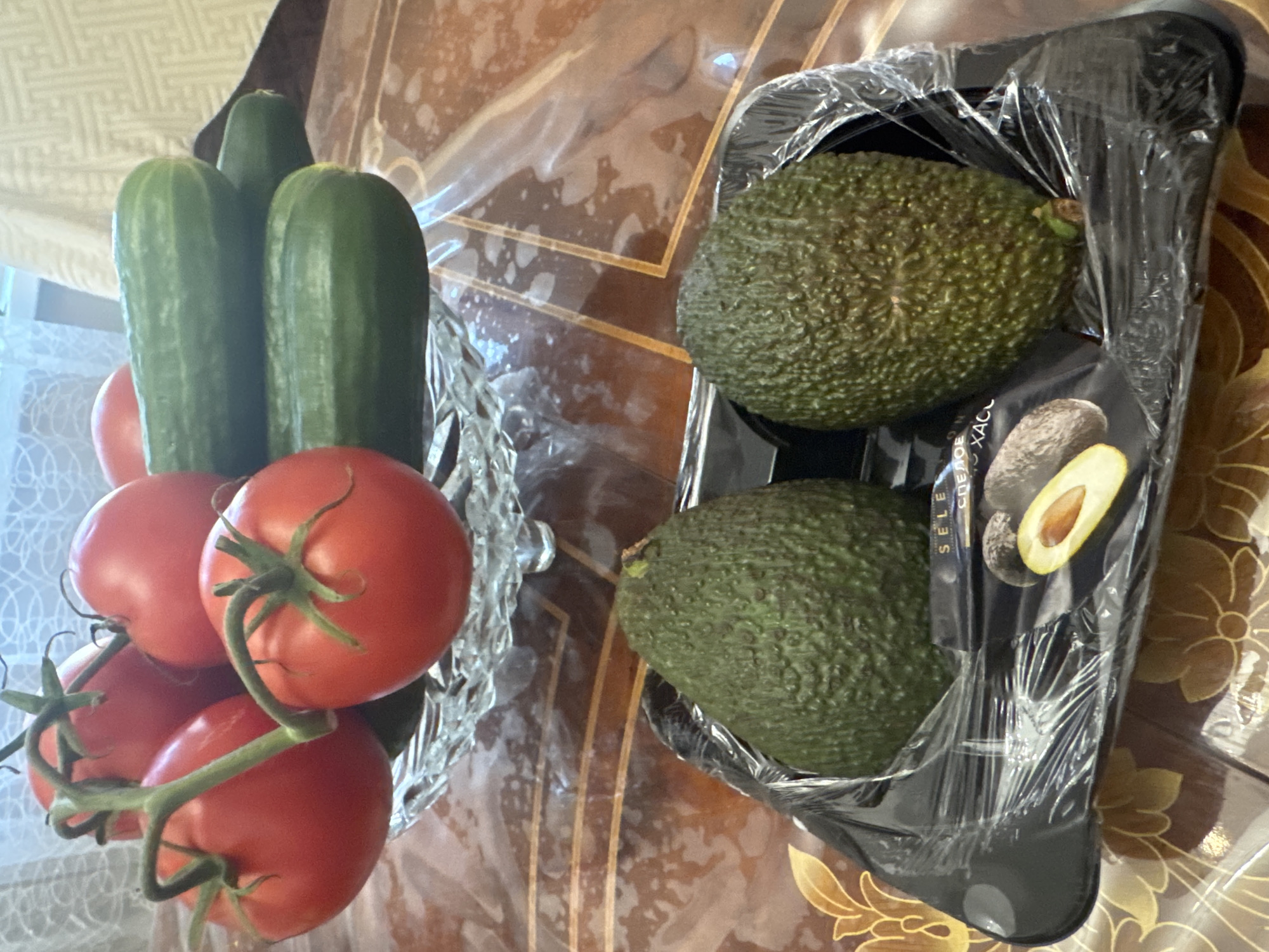 A close-up, eye-level shot shows a collection of fresh produce arranged on a table. In the foreground, two avocados are nestled in a black plastic tray covered with clear plastic wrap. The avocados are dark green and textured. To the left of the tray, a small image of a halved avocado is visible on the packaging. In the background, a glass bowl holds several ripe red tomatoes with green stems. Behind the tomatoes, three green cucumbers are stacked. The table surface is a brown wood-grain laminate with a decorative pattern. The overall lighting is bright and natural, suggesting an indoor setting, possibly a kitchen or dining area. There are no people or animals present. The location is described as a table in Novosibirsk, Russia.