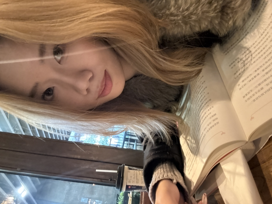 A young East Asian female with long, blonde-dyed hair and a slight smile is captured in a close-up, chest-up portrait. She wears a dark jacket with a grey faux fur collar over a grey knit sweater. Her right hand, with manicured nails, is visible near an open paperback book resting on a wooden table. The book displays text in Chinese characters, and a white napkin or paper is tucked into its left pages. To the left, a disposable beverage cup with a lid and a white straw is present, featuring the number "70107" and additional Chinese text. Behind the subject, a large window reveals an outdoor environment with green foliage, a metal railing, and hints of distant structures. The setting is an indoor establishment, consistent with a cafe or study area, located in Yuzhong District, China.