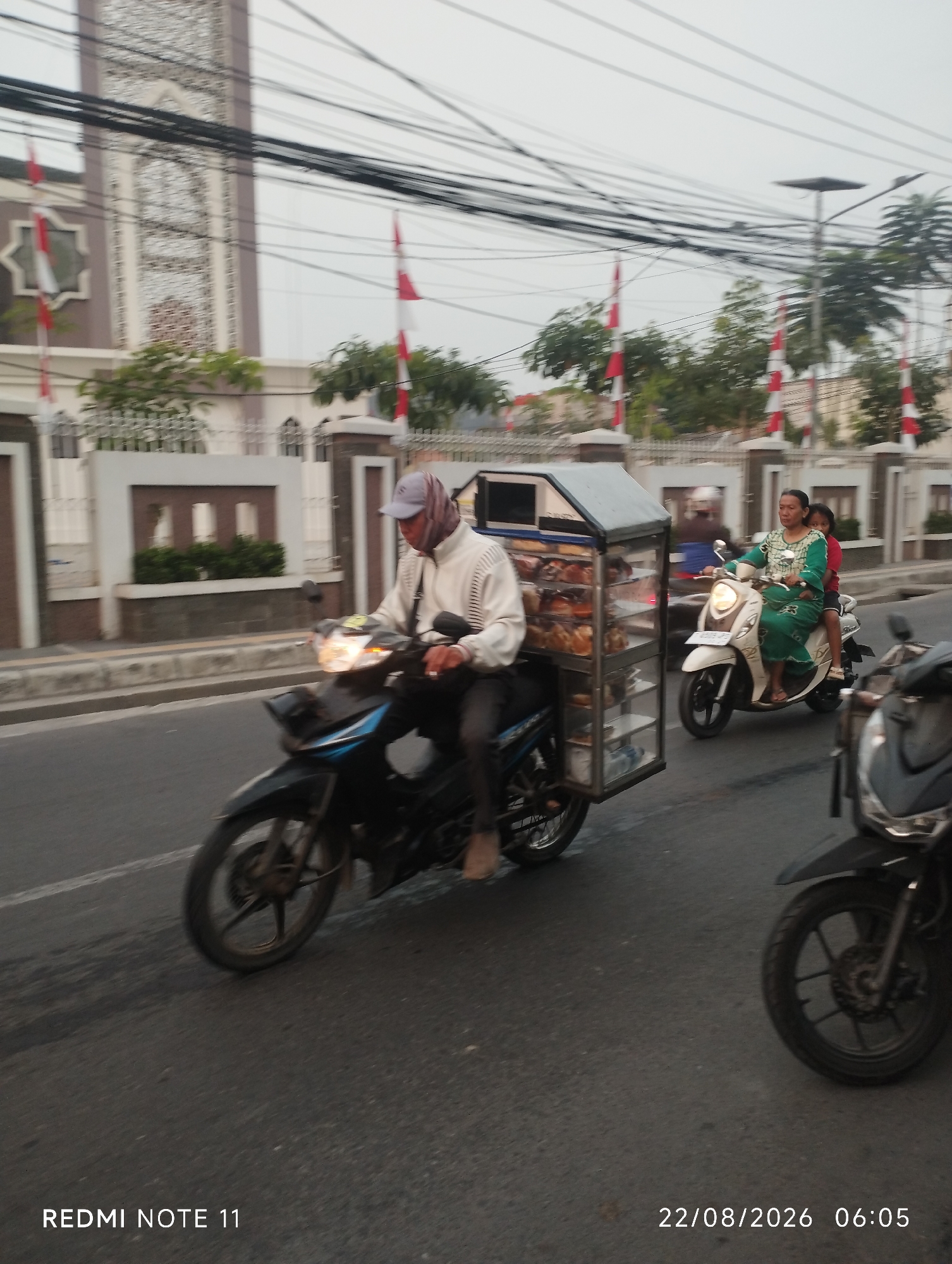 The image depicts a street scene in Karawang, Indonesia, on August 22, 2026, at 06:05. In the foreground, a man wearing a white jacket and a grey cap rides a black motorcycle, pulling a mobile food cart filled with baked goods. To his right, a woman in a green dress and a young girl in a red shirt ride a white scooter. In the background, a large building with Islamic architectural elements, possibly a mosque, stands with flags displayed. The street is paved, and there are power lines overhead. The overall lighting suggests early morning.