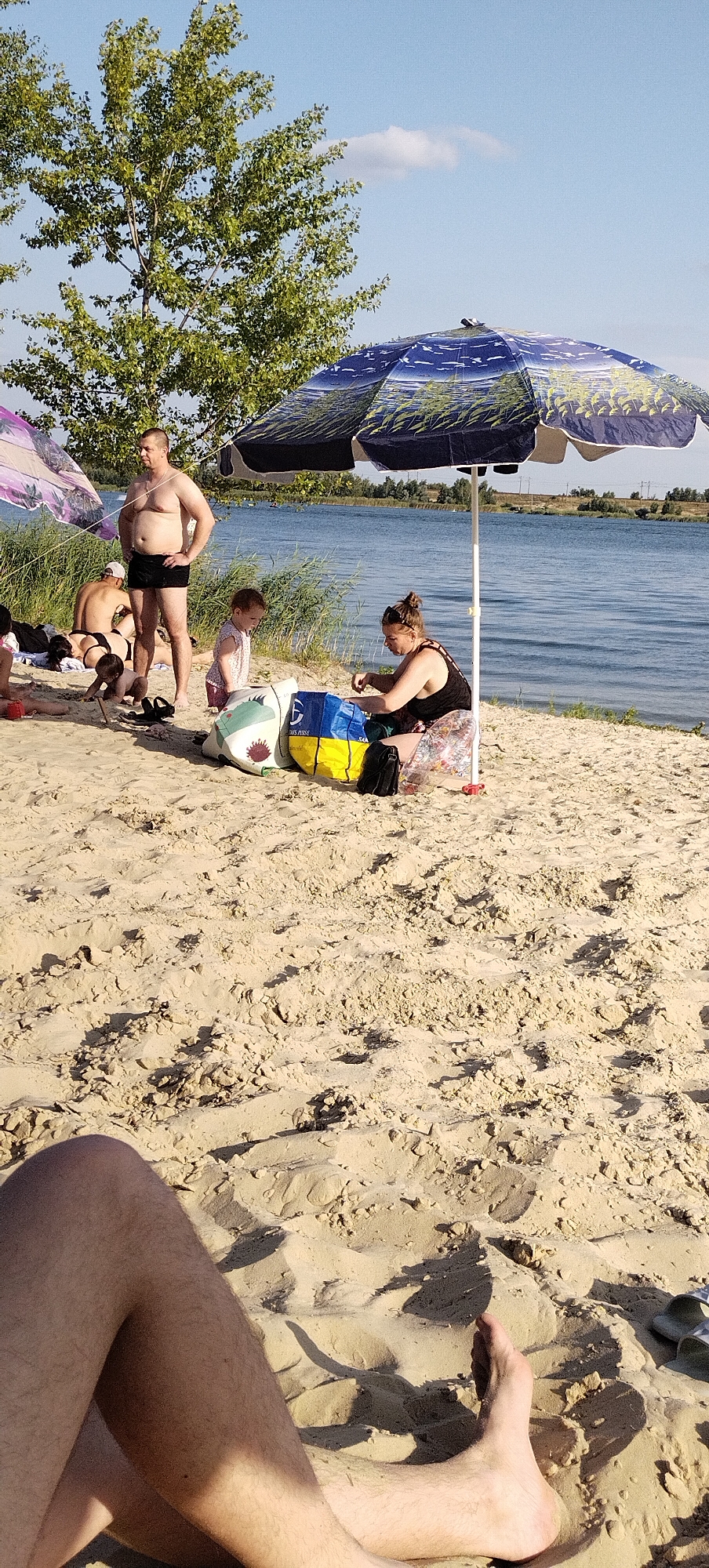 The image depicts a sandy beach scene with several people and objects. In the foreground, a person's legs and feet are visible, resting on the sand. Moving further into the image, a group of people are gathered on the beach. A man in black swim trunks stands in the middle, looking to his left. To his left, a couple is lying on the sand. A young child stands near the man, looking down. To the right, a woman in a black tank top and a patterned skirt is seated under a large umbrella with a blue and white pattern. In front of her are two bags, one with a shark design and another with blue and yellow panels. Behind the people, a body of water, likely a lake or river, is visible with vegetation lining its banks. The sky is blue with a few scattered clouds. The overall impression is a relaxed day at a recreational area. This scene is consistent with a location in the city of Bezliudivka, Ukraine, known for its beaches and recreational areas along the Bezliudivka reservoir.