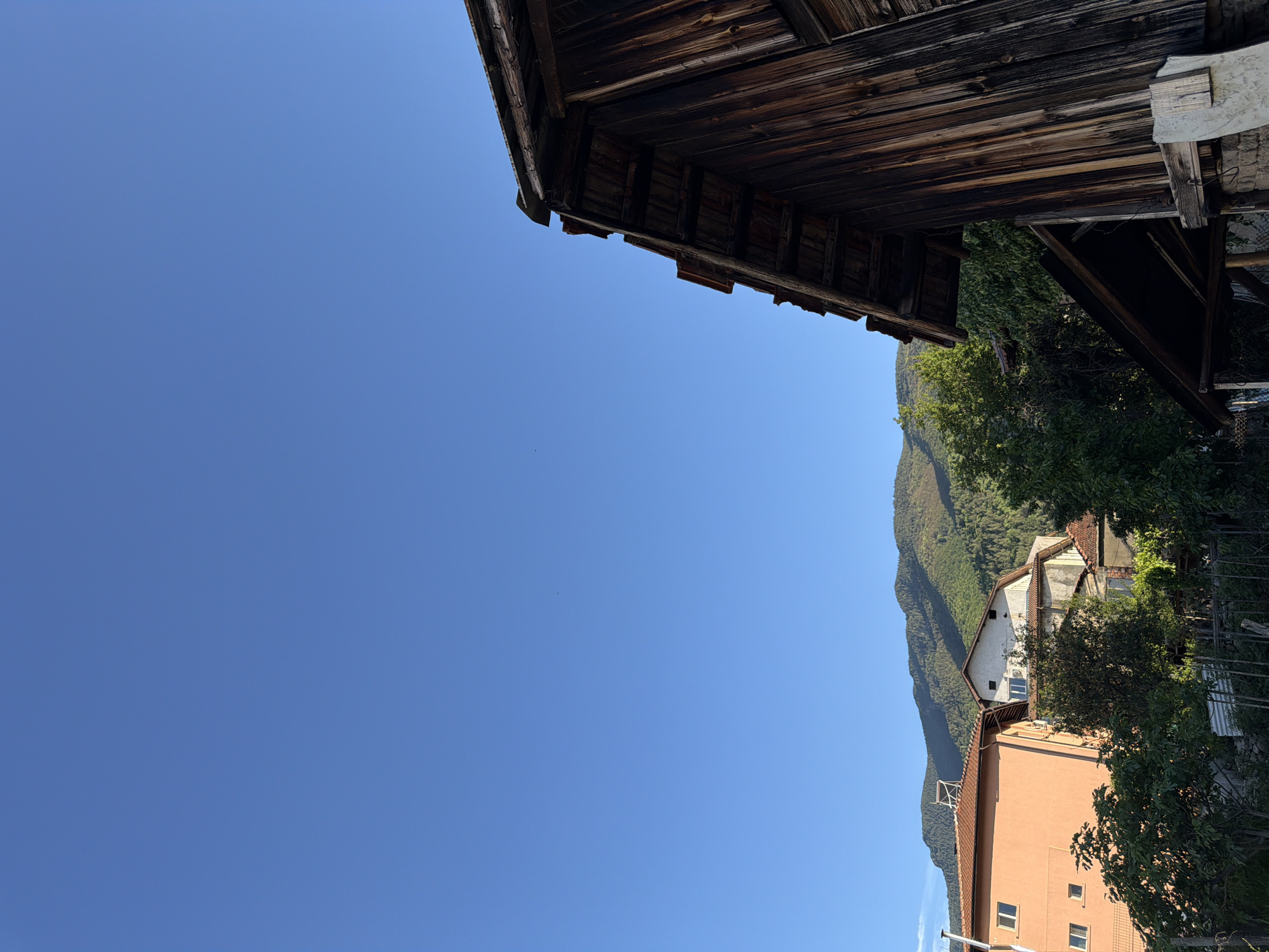 The image shows a view in Sapareva Banya, Bulgaria, with a predominantly clear blue sky. In the upper part of the frame is the corner of a weathered wooden structure, possibly a roof or wall. Below that, a landscape featuring a mountain range covered in trees is visible, along with buildings nestled in the foliage. One building is peach-colored with windows, and another is white with a red roof. Trees partially obscure the buildings.