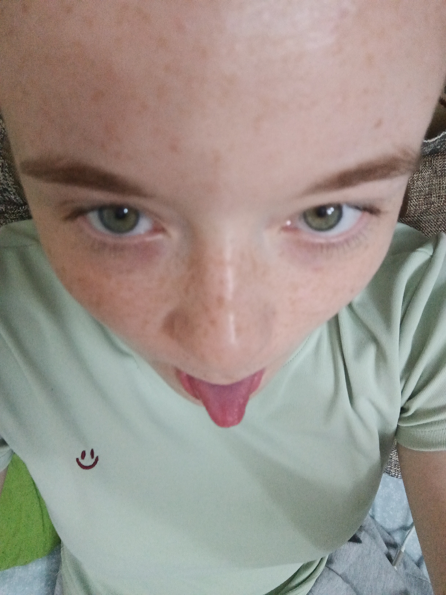 A close-up, overhead shot captures a young person with freckled skin sticking out their tongue. The subject's head is tilted back, with their eyes looking up towards the camera. Their left eye, with a green iris and dark pupil, is clearly visible, while the right eye is partially obscured. Thick, dark eyebrows frame their eyes. Their nose is prominent, and their mouth is open with a pink tongue extended. The subject is wearing a light green t-shirt with a small, dark red embroidered smiley face on the left side of the chest. The background is blurred and appears to be a patterned fabric, possibly a couch or blanket, with hints of green and gray fabric visible beneath. The lighting is bright, suggesting it is daytime. There is no visible text other than the smiley face on the t-shirt.