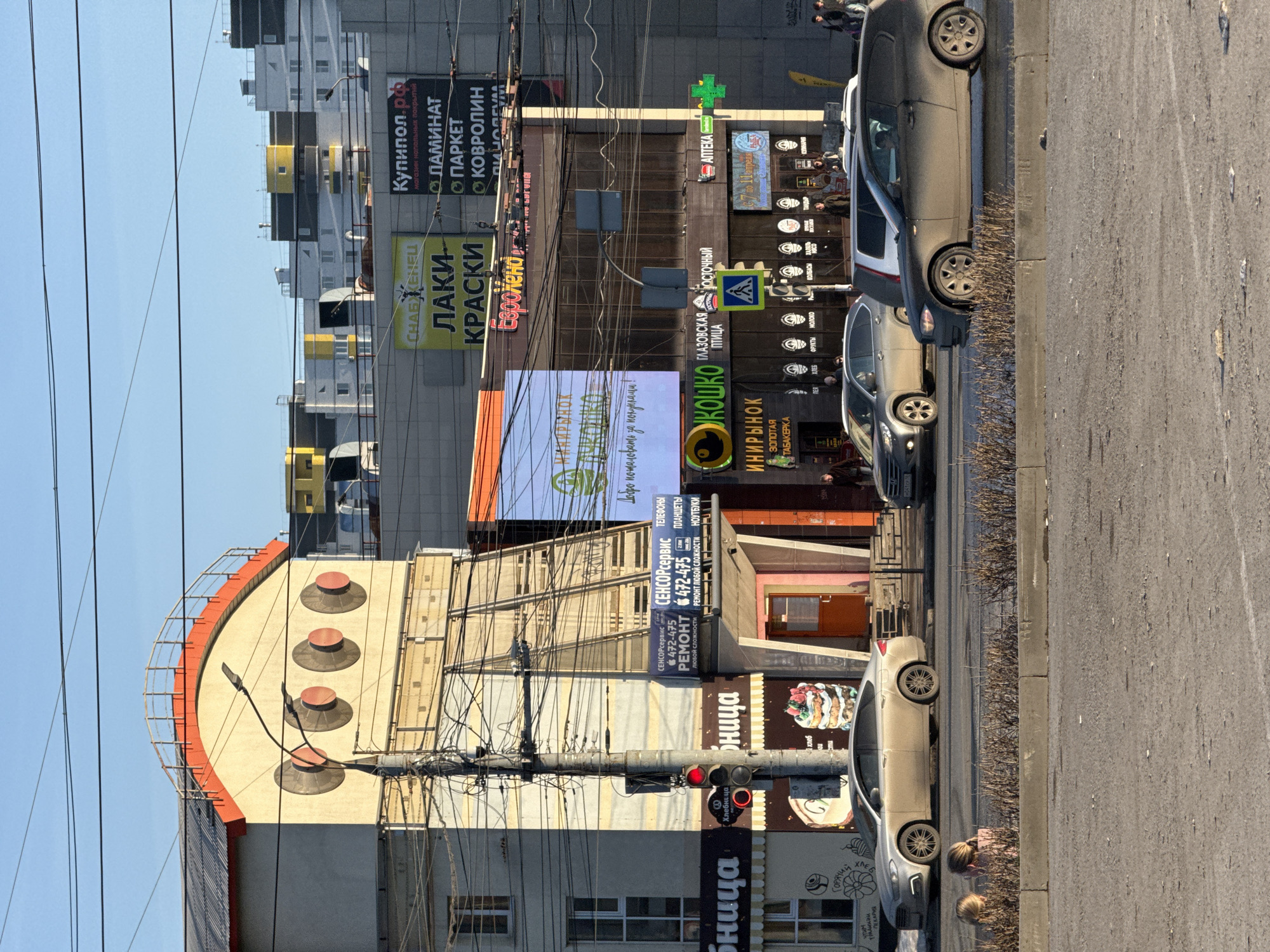 The image depicts a street scene in what is likely Izhevsk, Russia, given the Cyrillic lettering on the signs. The image shows a row of buildings of varying styles and heights, with advertising signage visible on the facades. Signage is for businesses such as a paint store (СНАБЖЕНЕЦ ЛАКИ КРАСКИ), a flooring store (Купипол.рф), a repair service (СЕНСОРсервис), a bakery (Бница), a grocery store (КОШКО), and a pharmacy (ΑΠΤΕΚΑ). Overhead power lines are prominent, crisscrossing the sky. Several cars are parked along the curb, and there are glimpses of pedestrians. The street appears to be paved and in moderate condition, with some wear and tear. The sky is clear and blue.