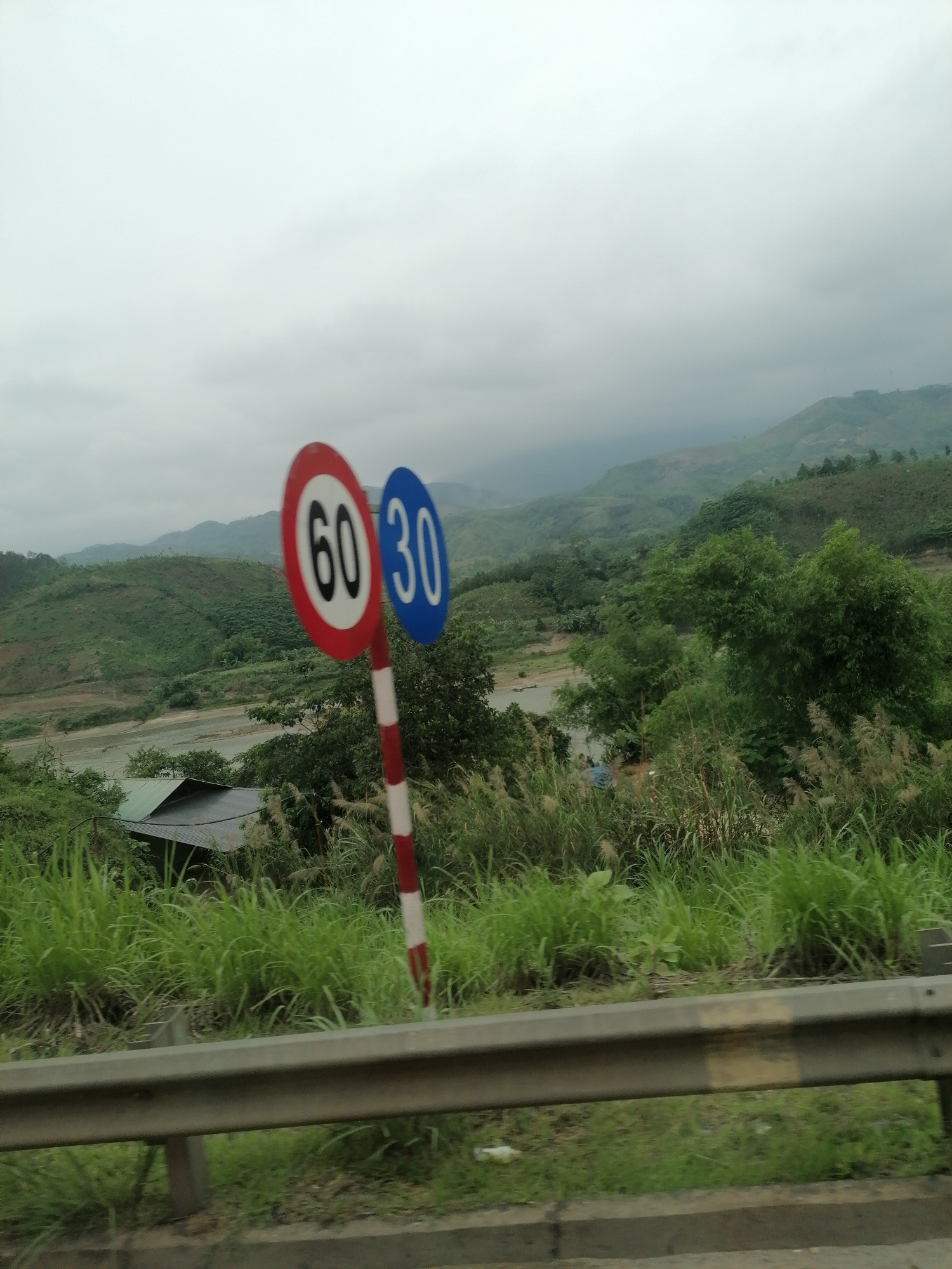 The image captures a scenic view from what appears to be a moving vehicle, looking out over a mountainous, verdant landscape under an overcast sky. In the immediate foreground, a metal guardrail runs horizontally across the bottom of the frame, hinting at a road alongside.
Dominating the foreground on the right side are two circular traffic signs attached to a single red and white striped pole. The first sign, closer to the viewer, is red with a white inner circle displaying the number "60" in black bold text, indicating a maximum speed limit. Behind and slightly to the right of this sign is a blue circular sign with white numbers "30," likely indicating a different speed regulation such as a minimum speed or a specific speed for certain conditions.
Beyond the signs and guardrail, lush green vegetation, including tall grasses and various trees and bushes, covers the slopes leading down towards a body of water, possibly a river or a wide stream, which winds through the landscape. A small, dark-roofed building or structure is partially visible amidst the foliage near the water's edge in the mid-ground.
In the background, rolling green hills and mountains stretch into the distance, appearing hazy under the heavy, light grey clouds that dominate the sky. The overall impression is one of a remote, natural setting, likely in a tropical or subtropical region given the dense greenery. The light suggests it's daytime, but the overcast conditions create a soft, diffused illumination. There are no people visible in the image.