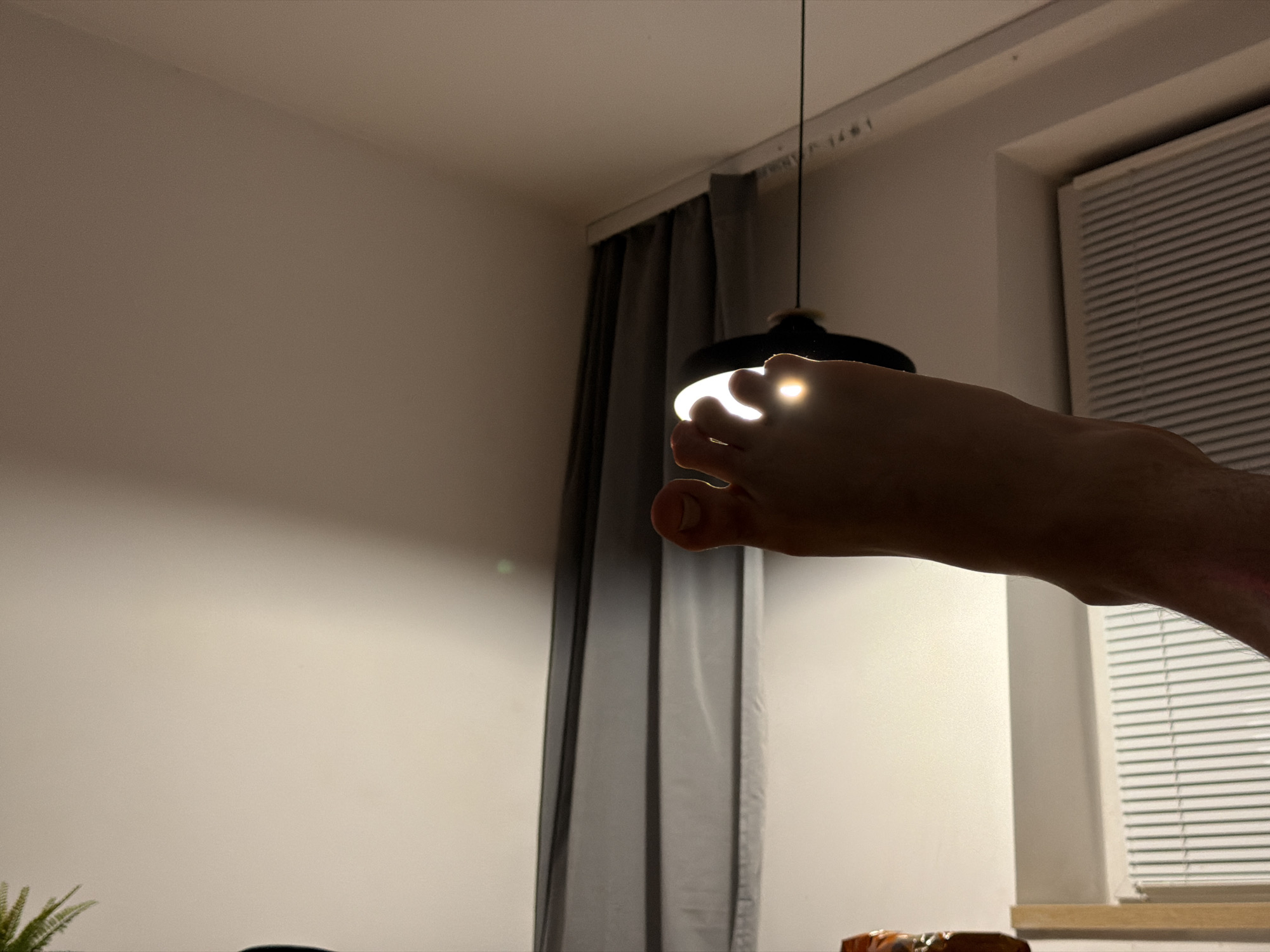 A person's foot is positioned in front of a black pendant light fixture, with the light shining through the spaces between the toes. The foot is the primary subject, silhouetted against the bright light. The background consists of a wall, a grey curtain, and a window with blinds. The scene appears to be indoors, possibly in a residential setting. There is no discernible indication of the city of Sosnowiec, Poland, or any specific urban environment.