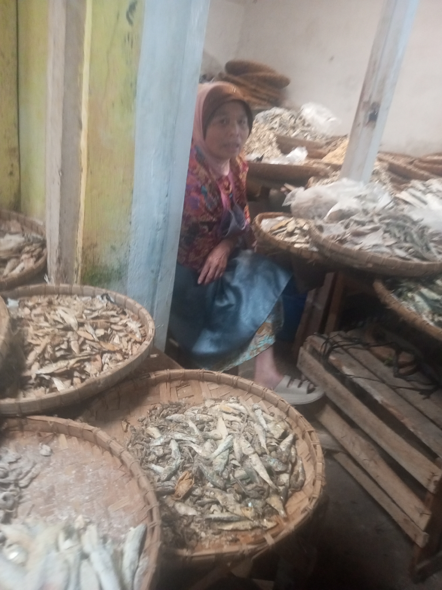 The image depicts a market stall in Kalibeber, Indonesia, where a woman wearing a headscarf and colorful patterned clothing is seated behind a display of dried fish. The fish are arranged in large, round woven baskets. The stall appears to be part of an indoor or covered market, with wooden beams and a rough wall visible in the background. The lighting is dim, creating shadows. In the foreground and to the left, other baskets filled with dried fish are partially visible. To the right, wooden crates are stacked. The woman's feet are visible, wearing light-colored shoes.