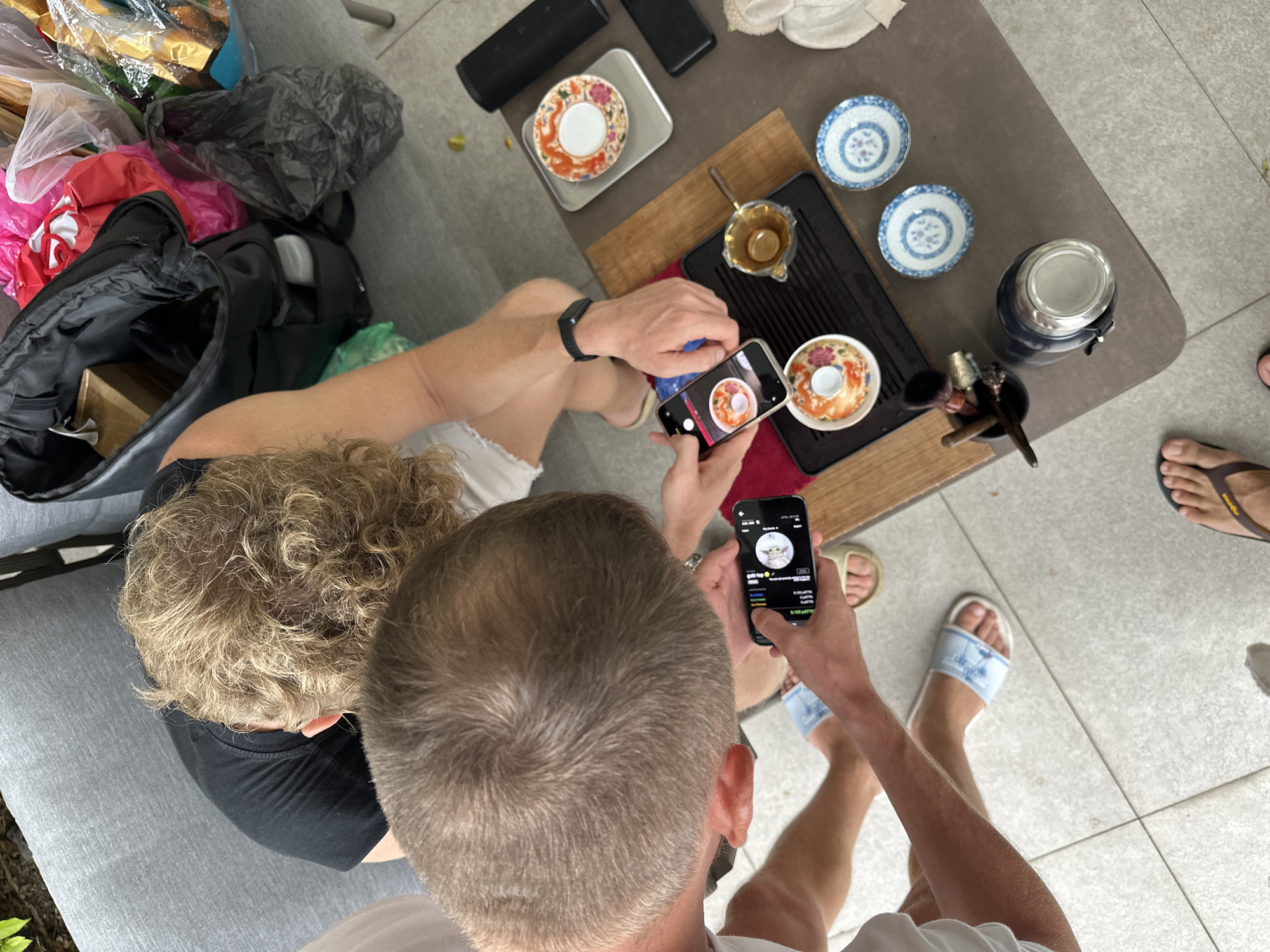 In an outdoor setting, two individuals are engaged in a tea ceremony. The scene is viewed from above, focusing on a table with tea-related items. One person, with short, light brown curly hair, wears a black t-shirt and is seated on a gray sofa. Another person, whose head and shoulders are visible, wears a white t-shirt and is partially obscured. A third person, whose legs and feet are visible, wears light blue slides with a palm tree pattern.
On the table, a black tea tray holds several ceramic pieces. Two small white and blue patterned plates are positioned to the right of the tray. A glass and metal container sits to the left of the plates, next to a small set of tea ceremony tools. A white and red patterned small bowl is on the tray, next to a clear glass pouring pitcher containing a golden liquid. Two mobile phones are being used: one is held by a person with their hands visible, capturing an image of the tea setup, and the other phone displays an image on its screen.
The ground surface is tiled with light gray squares. In the background, a black bag and various colored shopping bags are present on the sofa. The overall environment suggests a casual outdoor gathering.