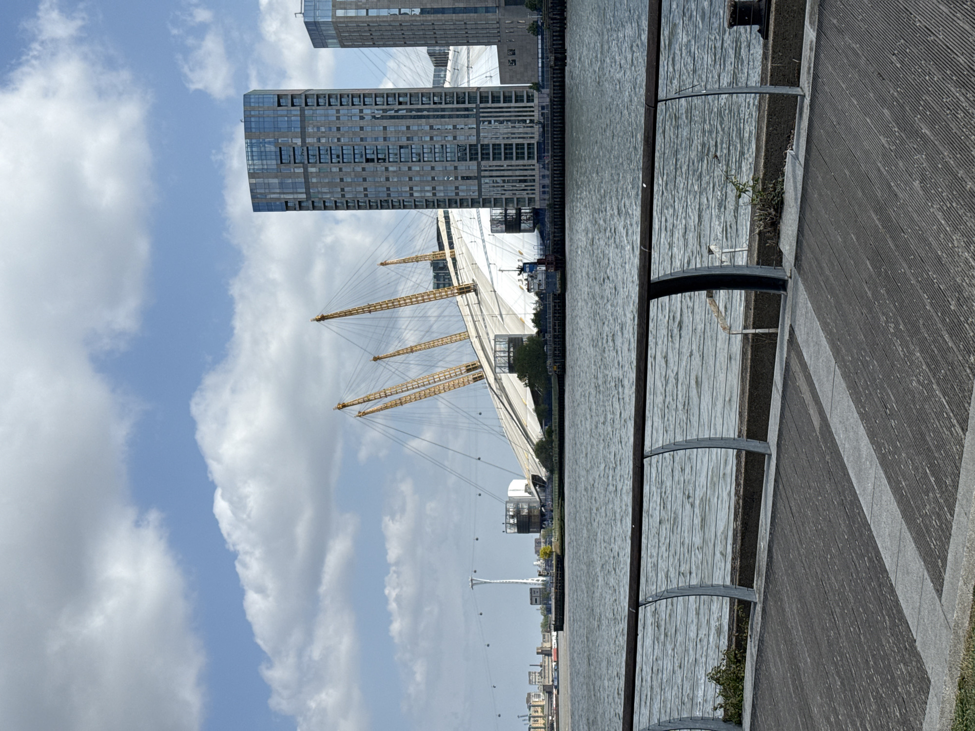 This outdoor image presents a modern urban waterside landscape in London, United Kingdom, prominently featuring the O2 Arena. The viewpoint is from a paved walkway or dock, looking across a body of water, likely the River Thames, towards the opposite bank.
In the mid-ground, the iconic white, tent-like structure of the O2 Arena stands out with its distinctive yellow support masts and intricate web of tension cables. To its right, a collection of contemporary high-rise buildings with reflective glass and concrete facades rises, showcasing modern urban development. Further in the background, a tall white pylon, identifiable as part of the IFS Cloud Cable Car system, is visible, with several spherical elements suspended along its cables.
The foreground on the right features a textured paved area, possibly made of wood or composite material, running parallel to the water. A metal railing or barrier separates this walkway from the rippled water body, which occupies the lower-left section of the frame. A small patch of green vegetation grows near the barrier.
The sky overhead is a bright blue, dotted with scattered white clouds, indicating fair weather conditions. The scene is illuminated by clear, natural daylight, suggesting it is daytime, likely late morning or early afternoon. No specific human activity or discernible text is visible within the frame.
