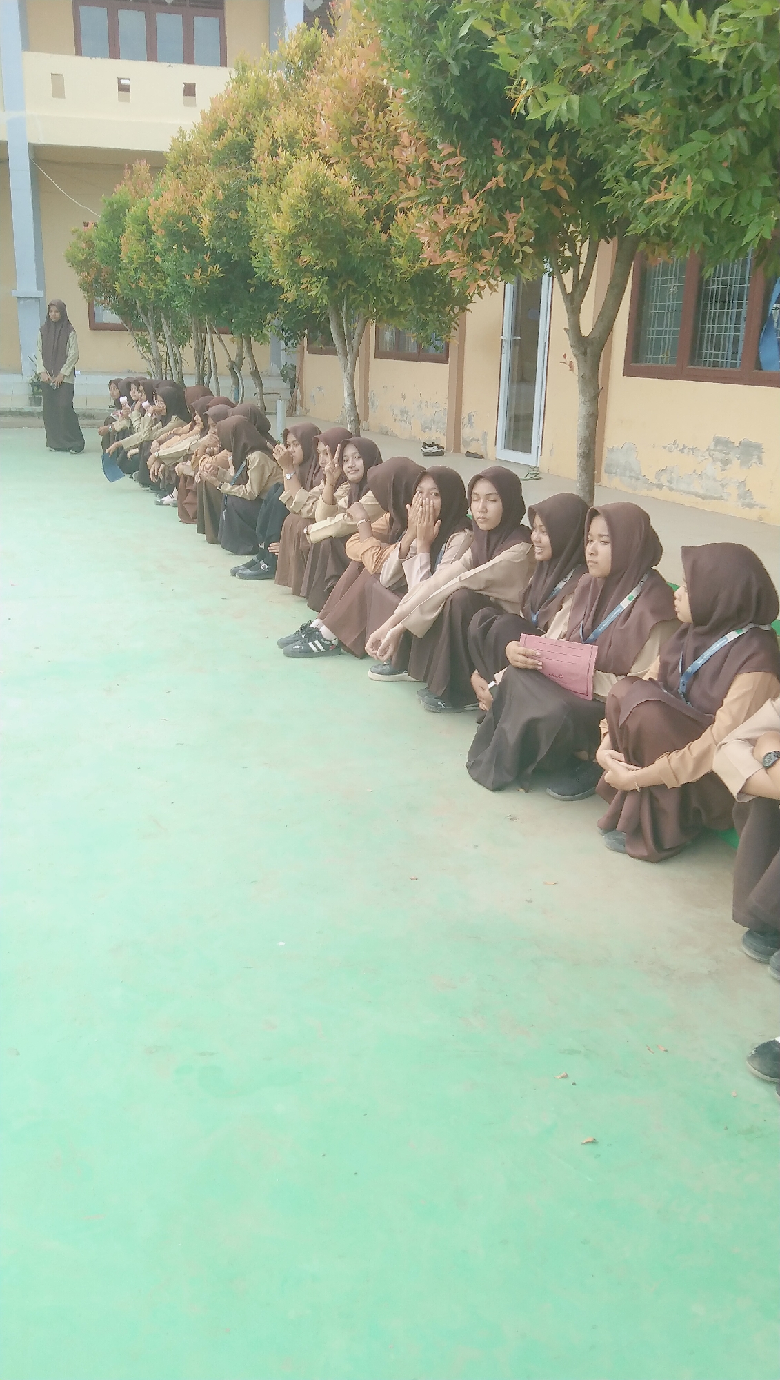 In the city of Rhieng Blang, Indonesia, a group of female students, dressed in brown hijabs and school uniforms, are seated in a single file line on a light green paved surface, likely a schoolyard or courtyard. The students are facing to the right of the frame, with their legs bent and feet tucked beneath them. Some students are looking forward, while others are looking down or at each other. One student in the foreground is holding a pink folder. Behind the students, a building with yellow walls and brown window frames is visible. Lush green trees with reddish-brown leaves are planted in a row between the students and the building. The overall atmosphere appears to be a casual gathering or waiting period during the school day.