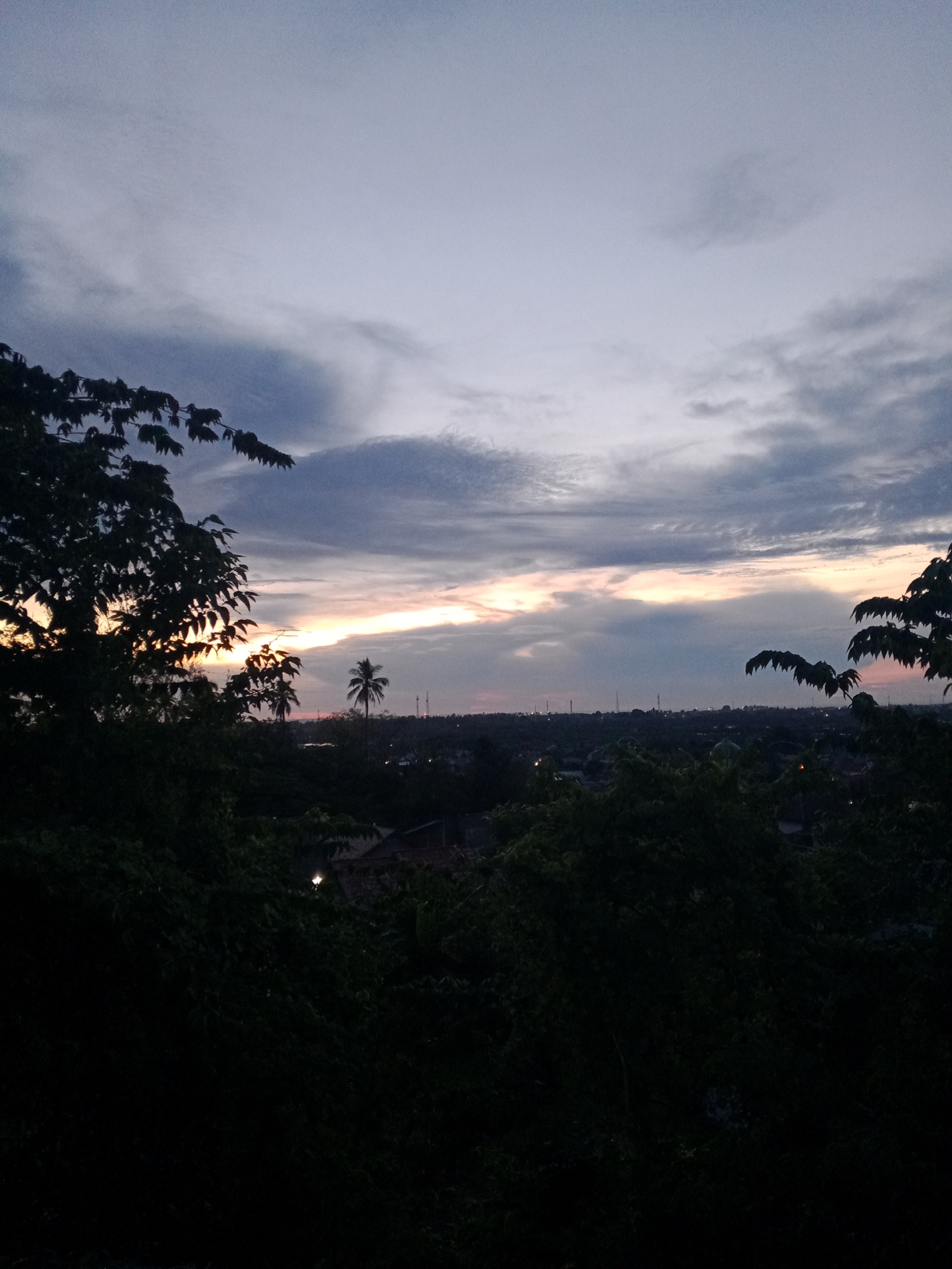 The image depicts a twilight or dusk scene in what appears to be a residential area, observed from an elevated perspective, potentially a hillside or building. In the middle ground, there are rooftops visible amongst dense trees. A single palm tree stands taller than the surrounding trees, silhouetted against the remaining daylight. Further back, there is a line of distant buildings or structures visible on the horizon line. Above, the sky is covered in clouds, with the last light of the setting sun shining through a gap, creating a yellowish-orange glow. The lower portion of the image is primarily dark, with the foreground trees represented in silhouette. Based on the information provided, this location is presumed to be in Lhokseumawe, Indonesia. There are no people present in the image.